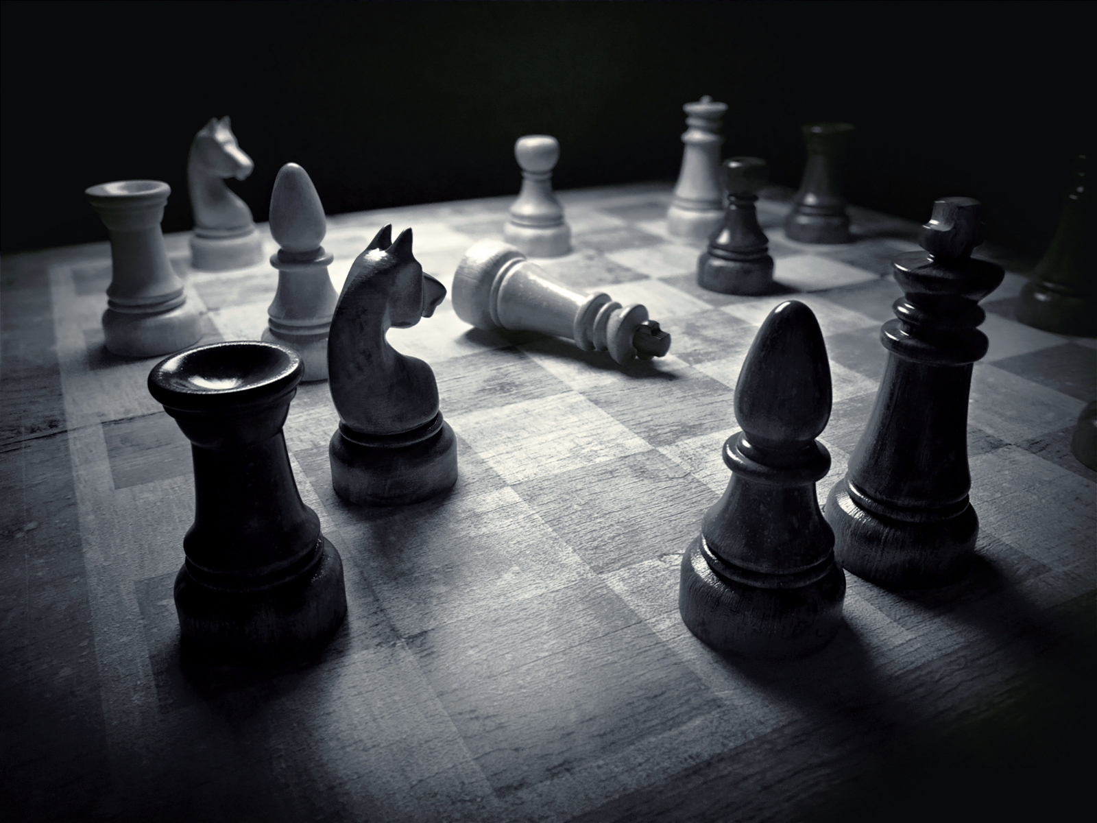 chess pieces on a chess board Background — Dramatic black and white photo of chess pieces on a worn wooden board with fallen pieces under low-key lighting.
