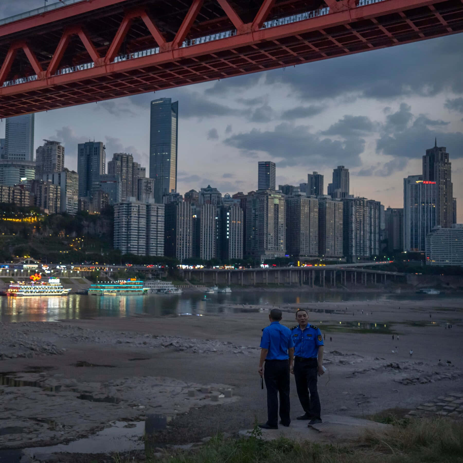 Two Police Officers Stand Under A Bridge In A City