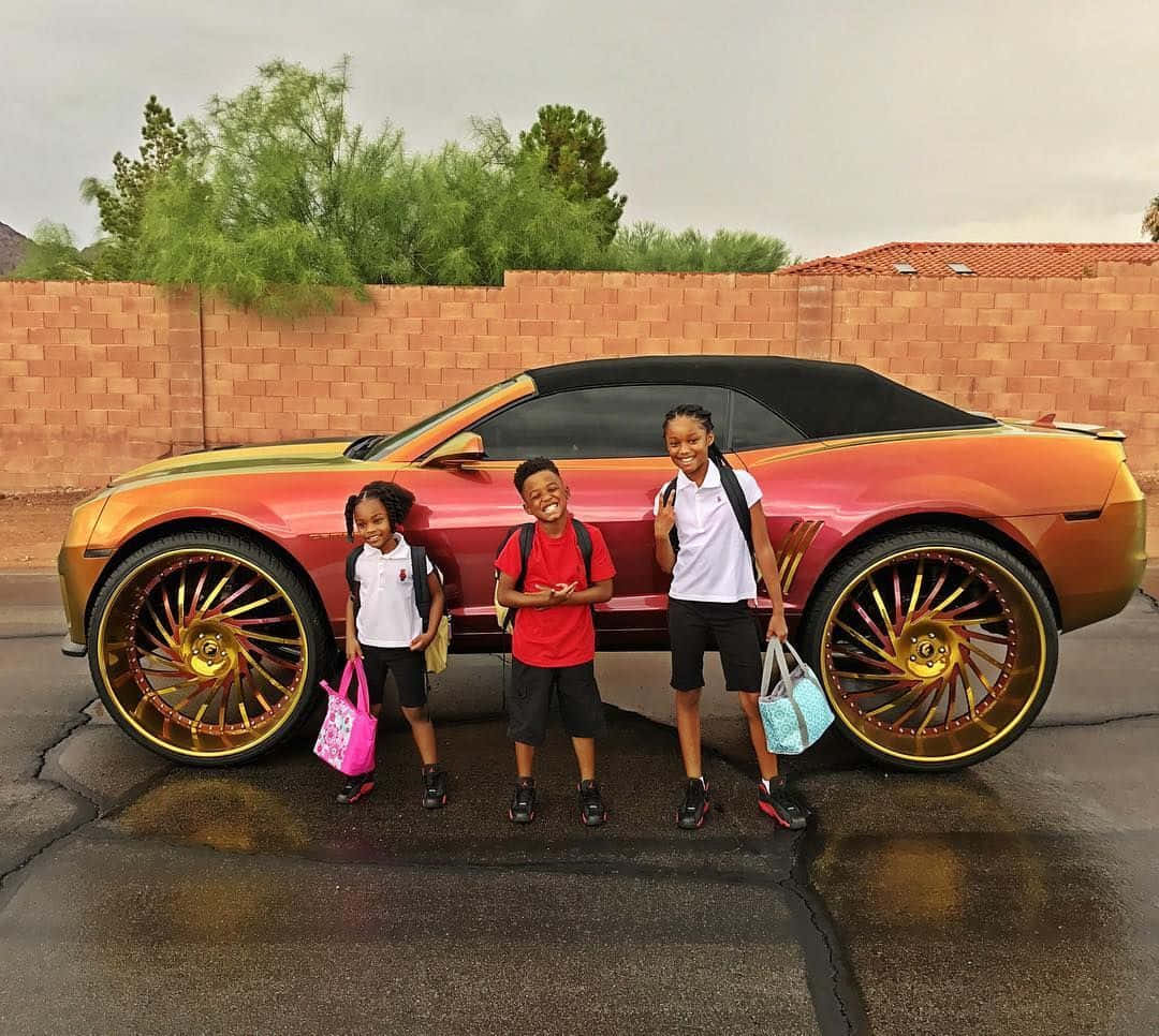 Tre børn stående ved siden af en bil med store hjul Wallpaper