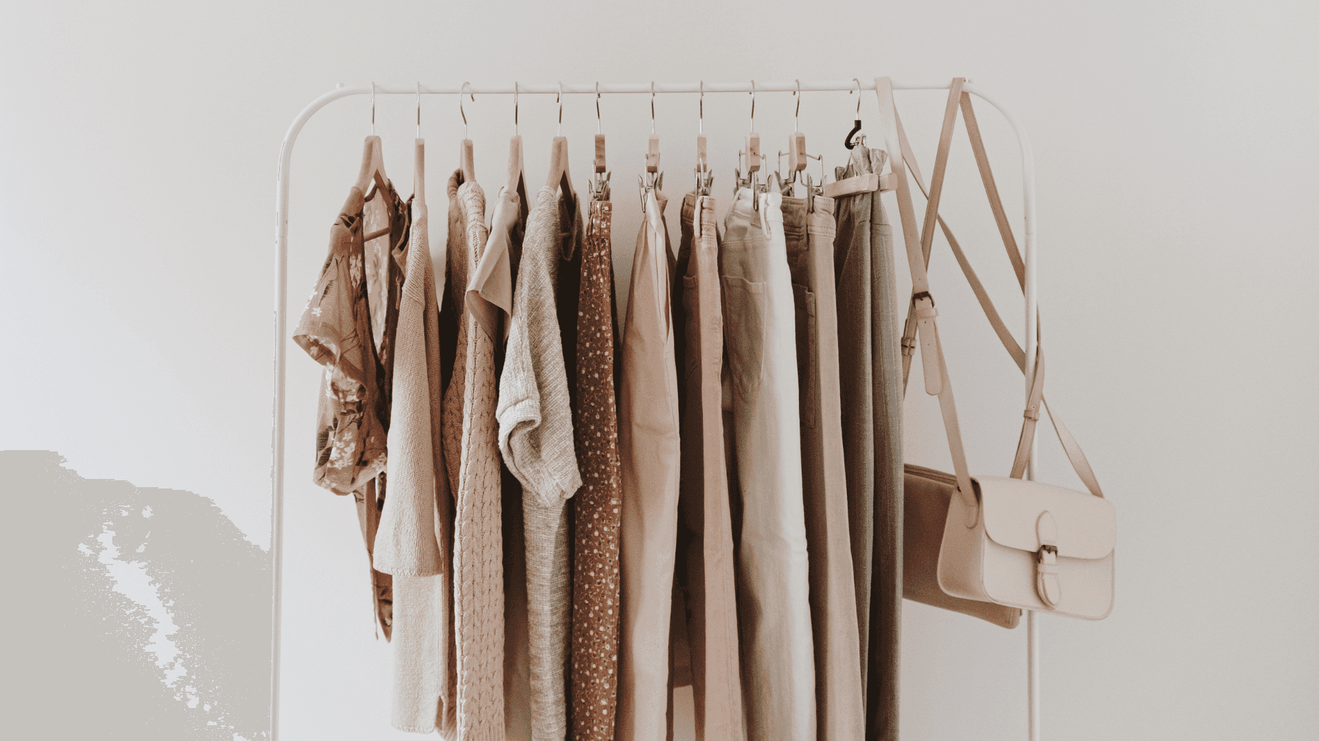 Update your wardrobe this summer with these stylish clothes Background — A minimalist clothing rack displays earth-toned garments and a beige handbag against a plain white wall.