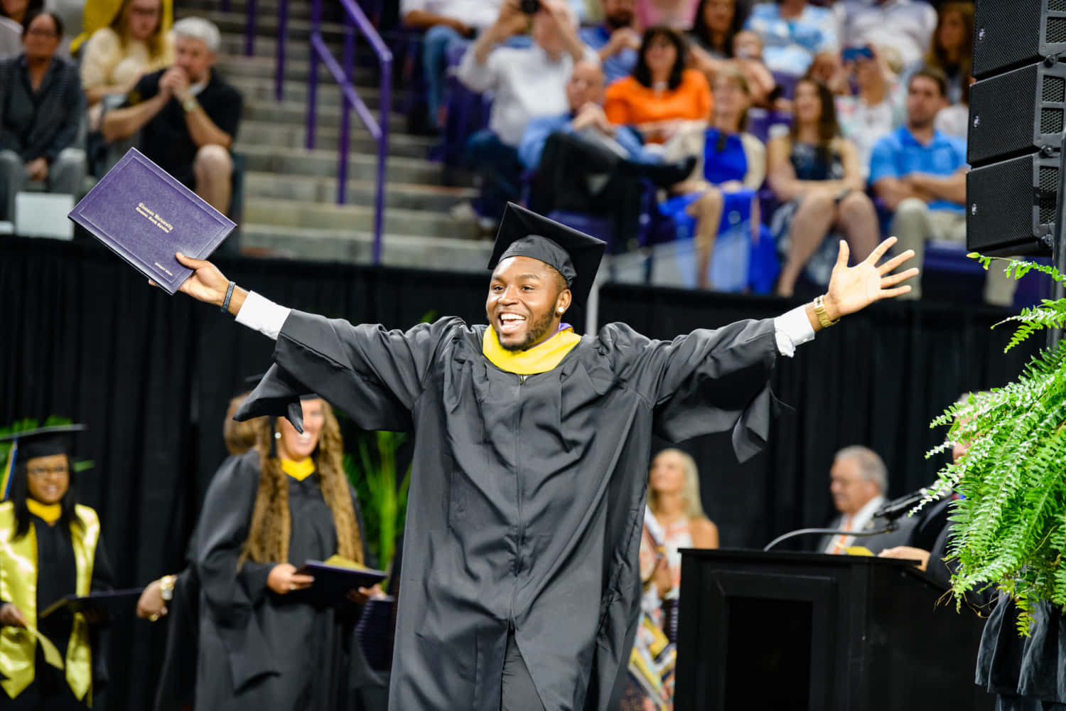 A Graduate In A Graduation Gown With His Arms Raised