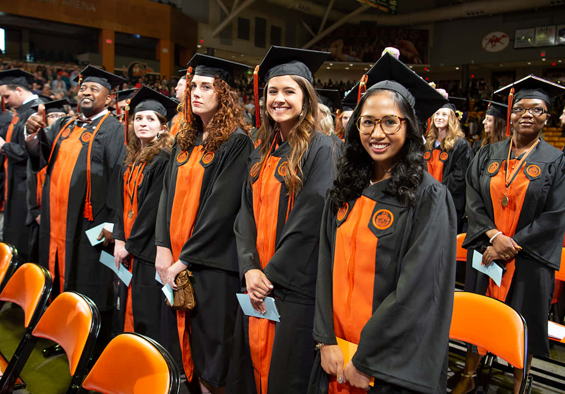 En gruppe af studerende i orange akademikerkapper stående i en stor auditorium