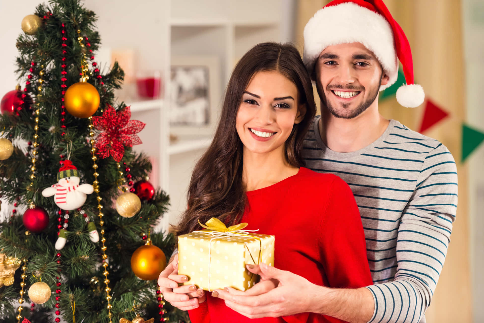 A Couple Holding A Christmas Gift In Front Of A Christmas Tree