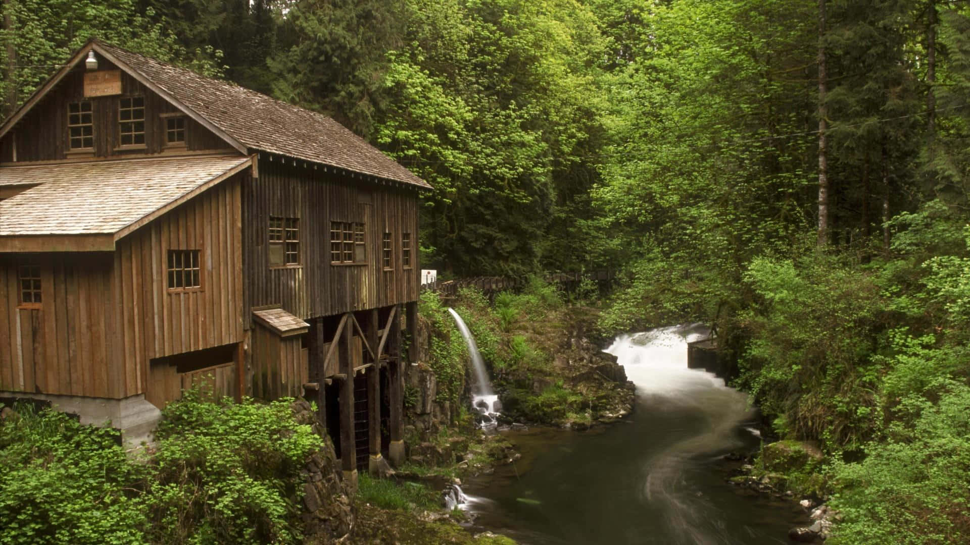 A Wooden Mill Is Surrounded By A River And Trees