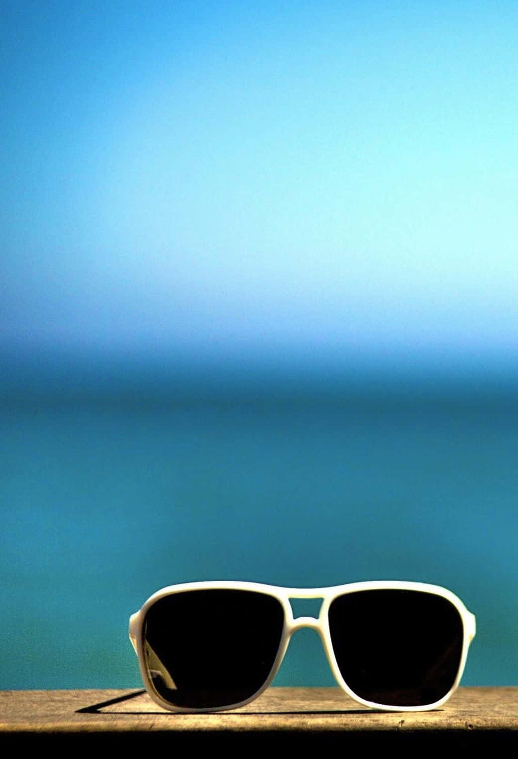 Sunglasses On A Wooden Table With The Ocean In The Background Wallpaper