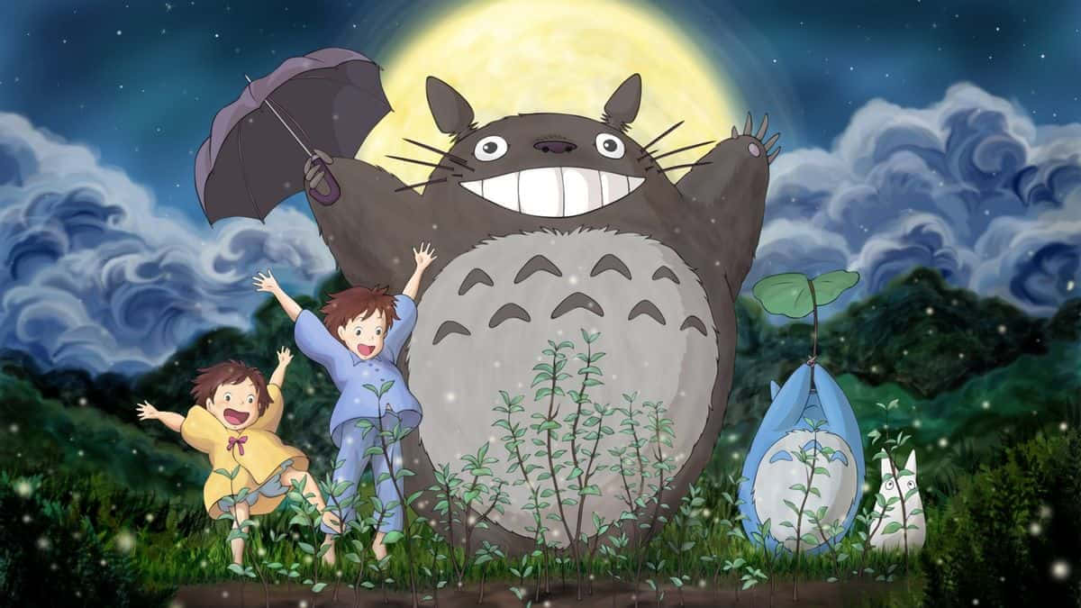 Engrupp Människor Står På Gräset Med Totoro Som Bakgrundsbild På Sin Dator Eller Mobiltelefon. Wallpaper