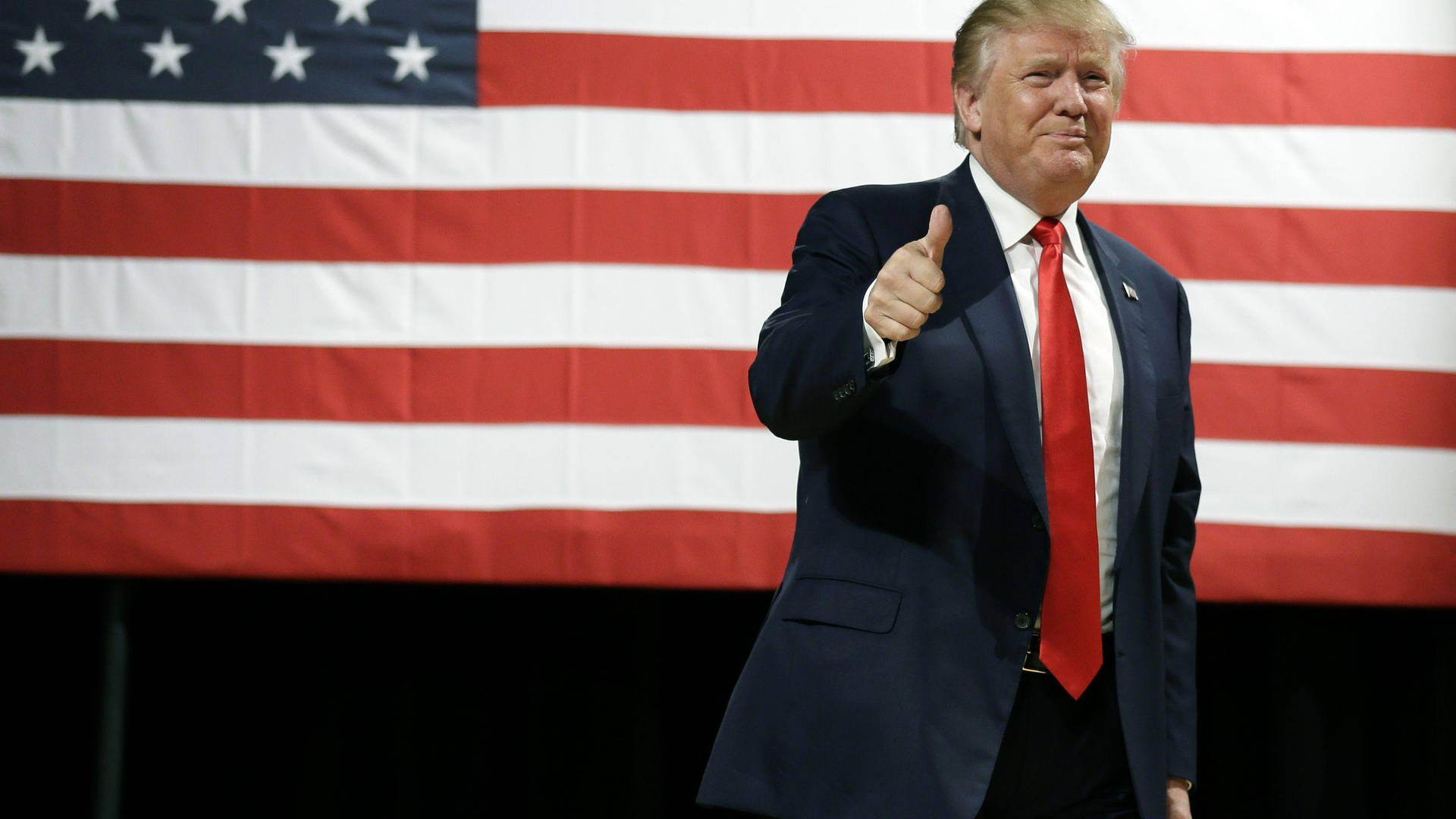 Donald Trump confidently raises his thumb in approval at the American flag. Wallpaper