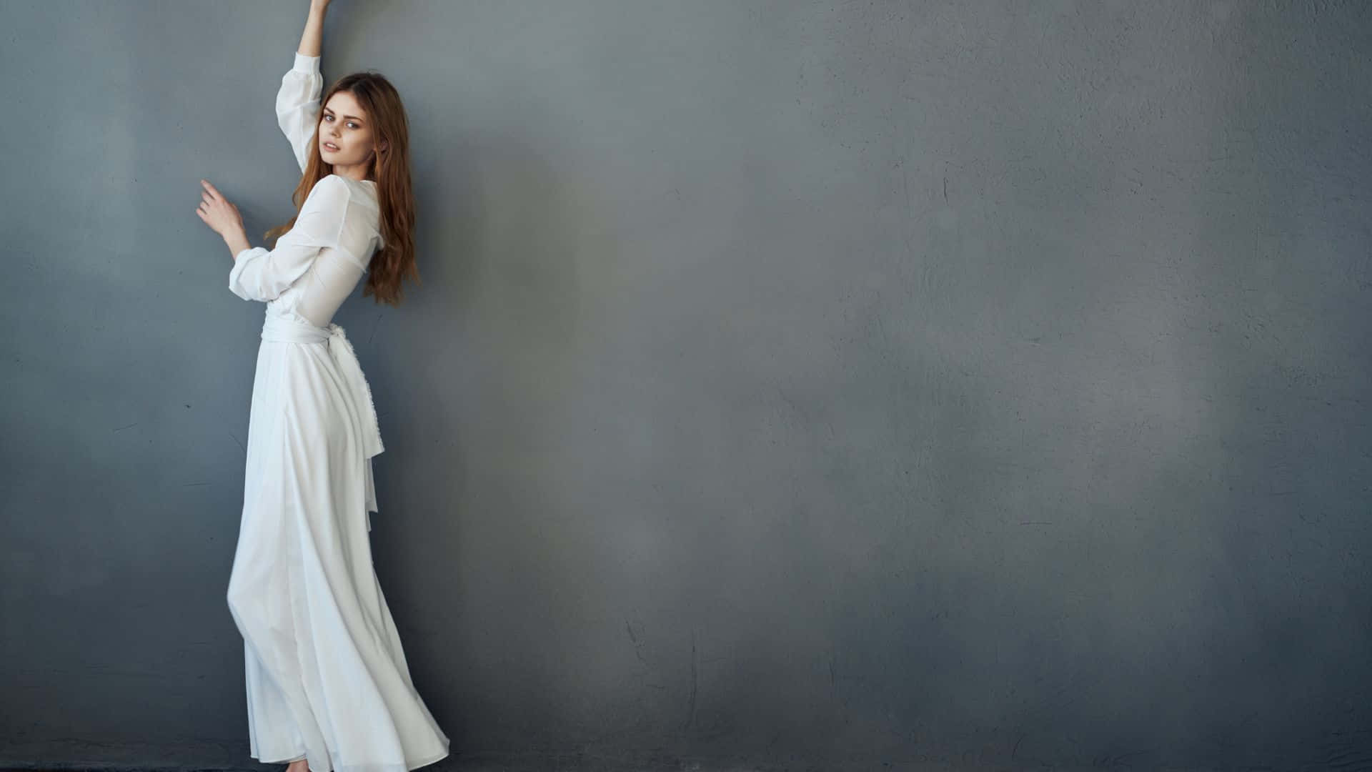 A Woman In A White Dress Leaning Against A Gray Wall