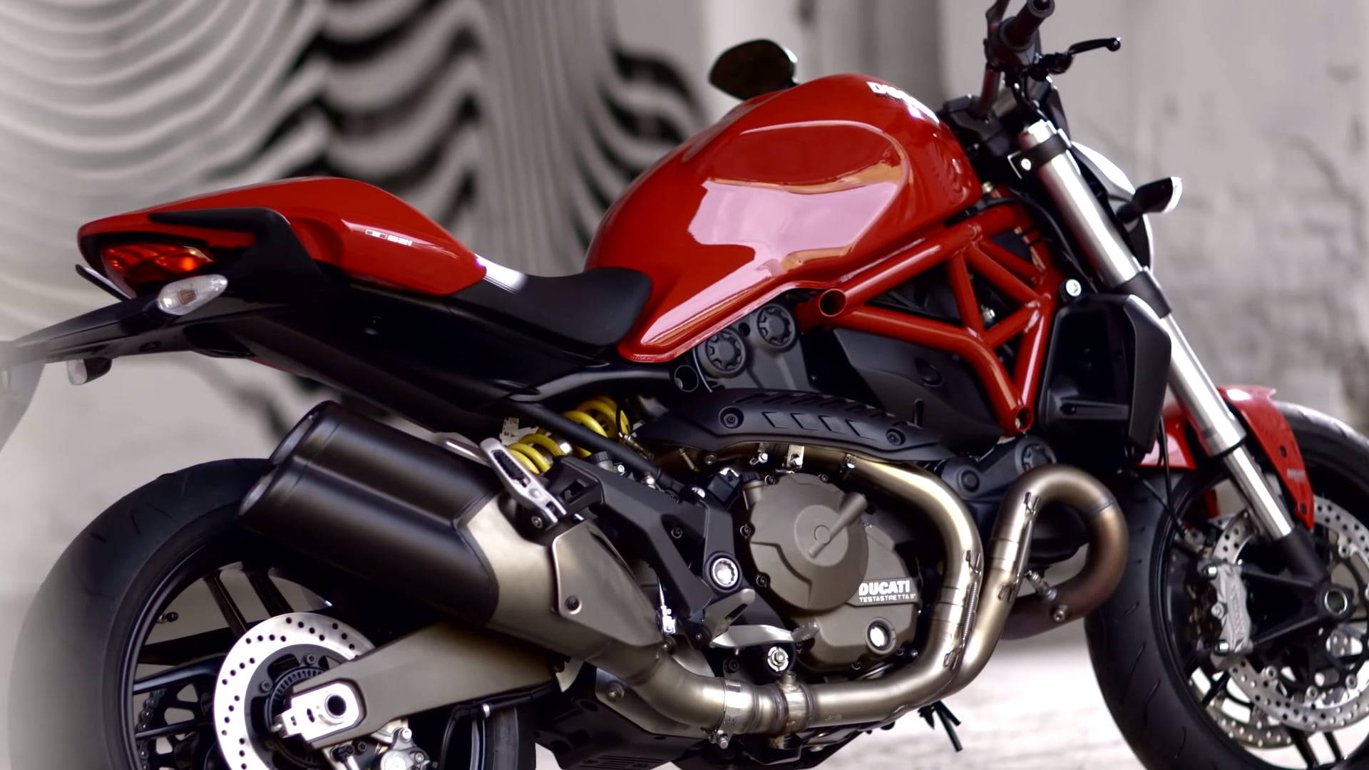 Ducati Monster 1200 - A High Killer Performance. Wallpaper — A glossy red Ducati motorcycle with exposed engine and chrome exhaust gleams against a wavy gray backdrop.