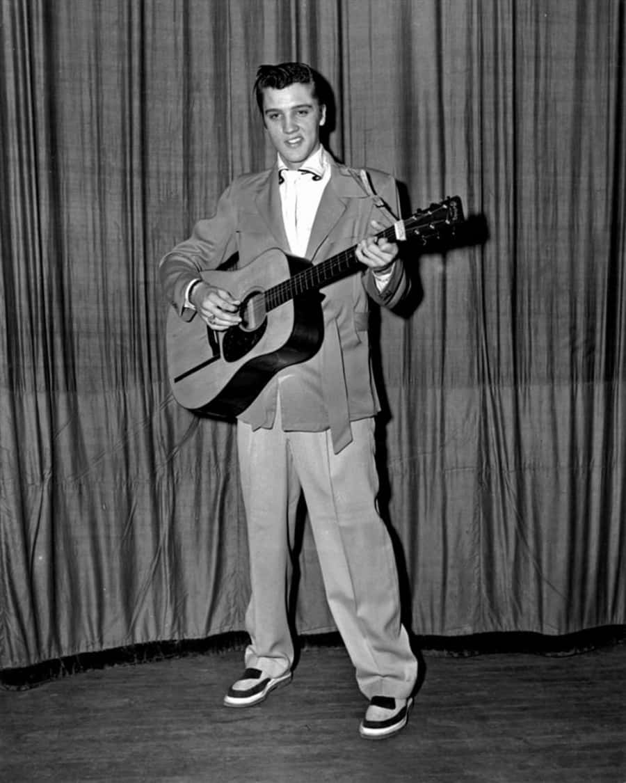 Elvispresley I En Kostym Och Slips Håller En Akustisk Gitarr.