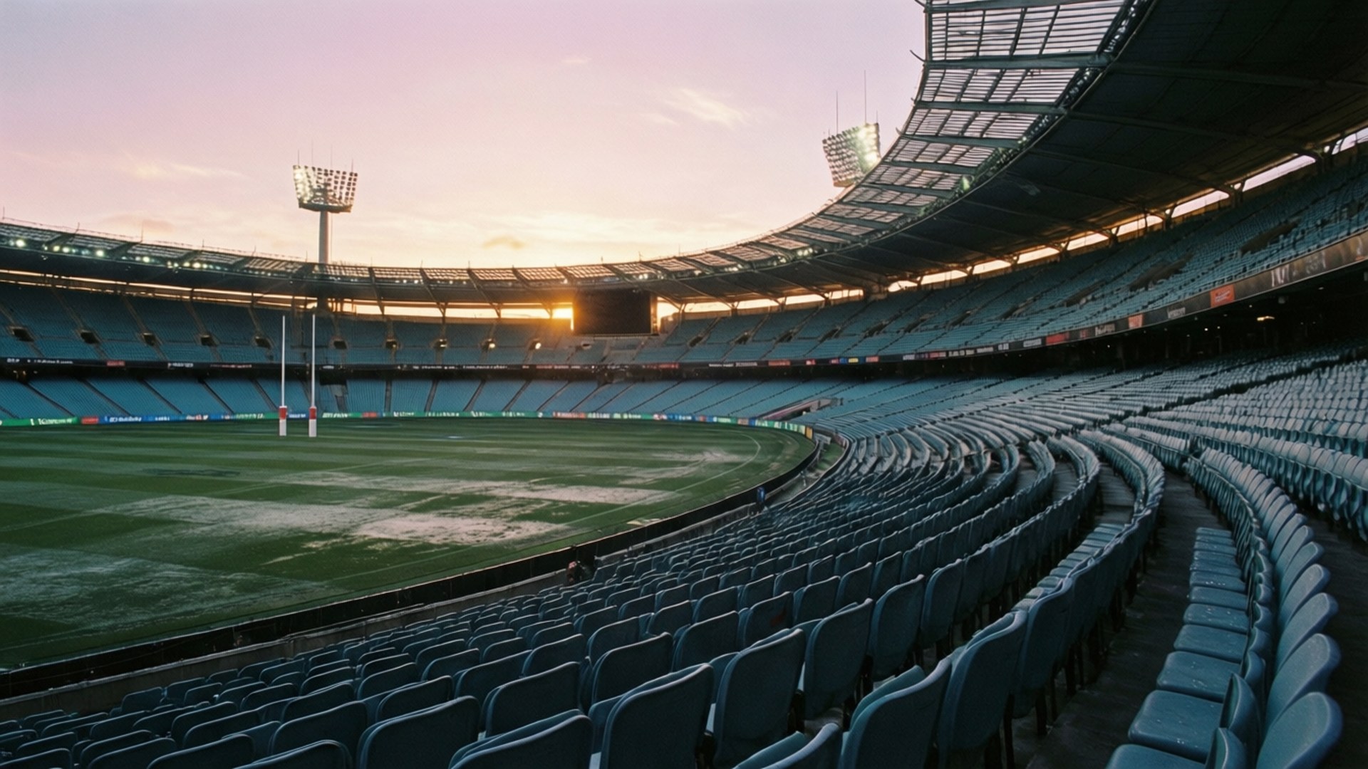 Empty Stadium Dawn Wallpaper — An empty stadium at sunset with rows of blue seats curving around a green field under a pink and orange sky.