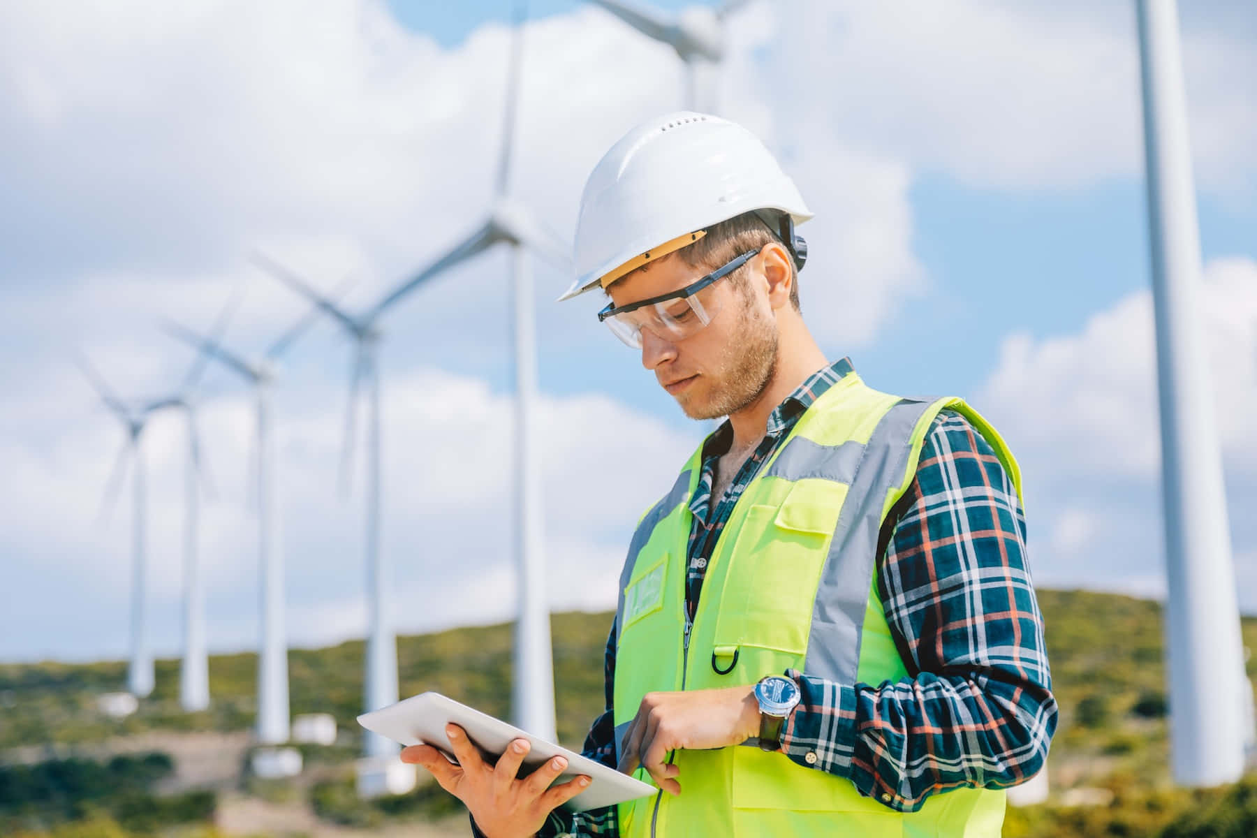 A Man In Hard Hat And Vest Is Using A Tablet In Front Of Wind Turbines