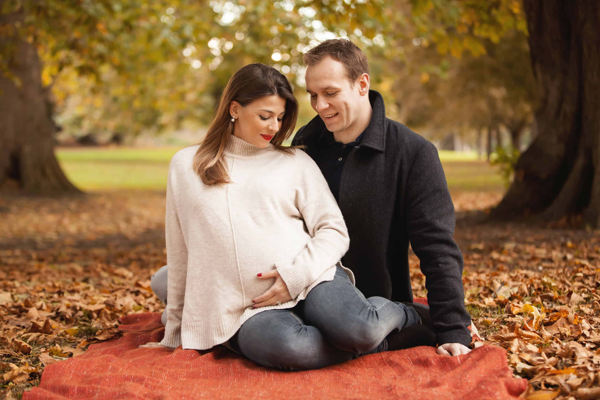 A Pregnant Couple Sitting On A Blanket In The Autumn Leaves