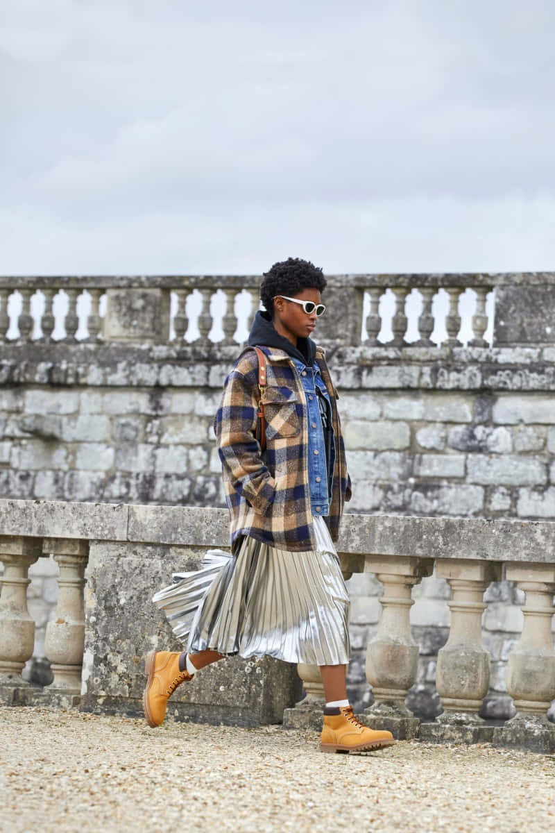 a woman in a plaid jacket and pleated skirt walking on a stone wall
