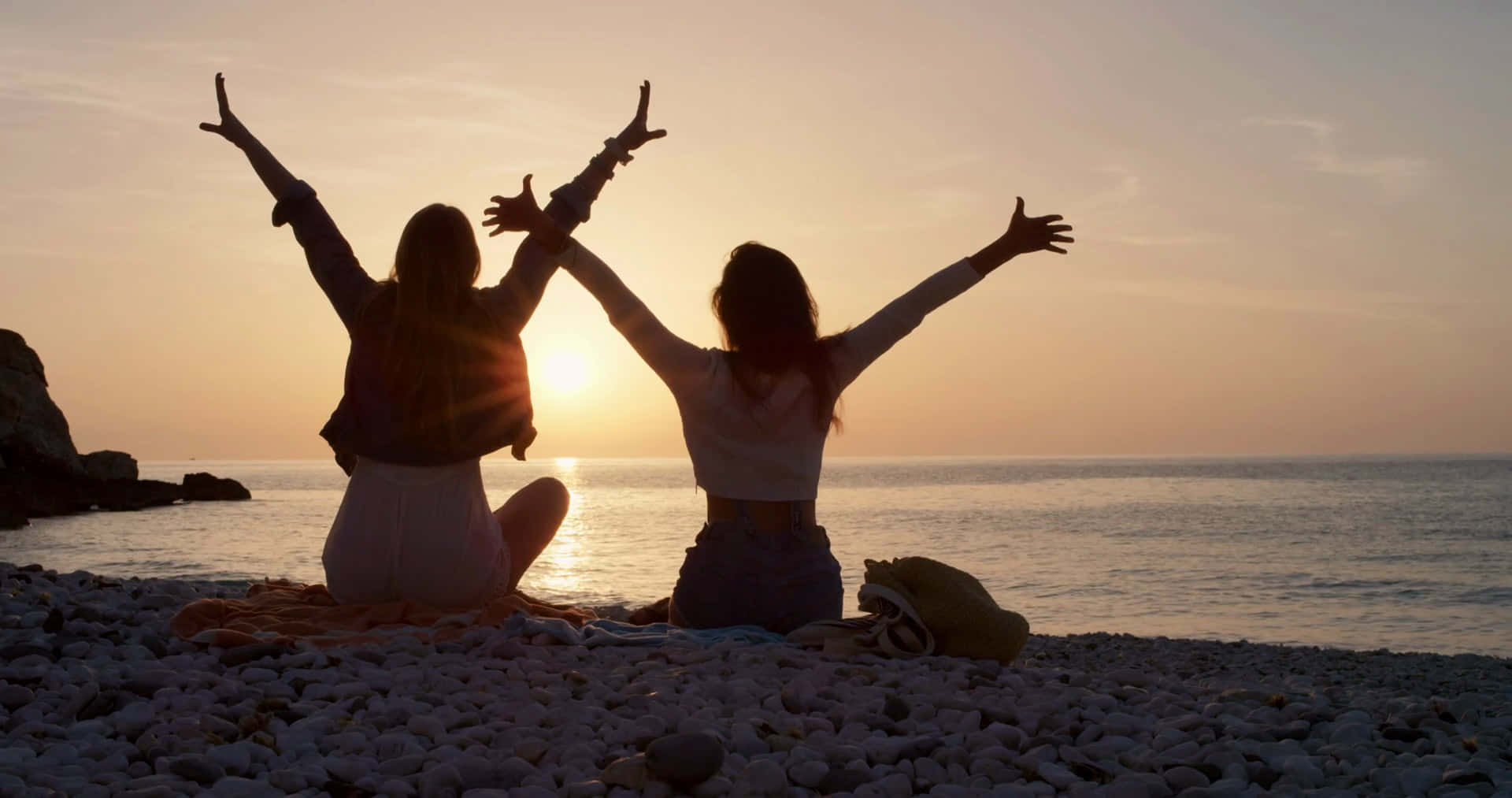 Two Women Sitting On The Beach With Their Hands Raised