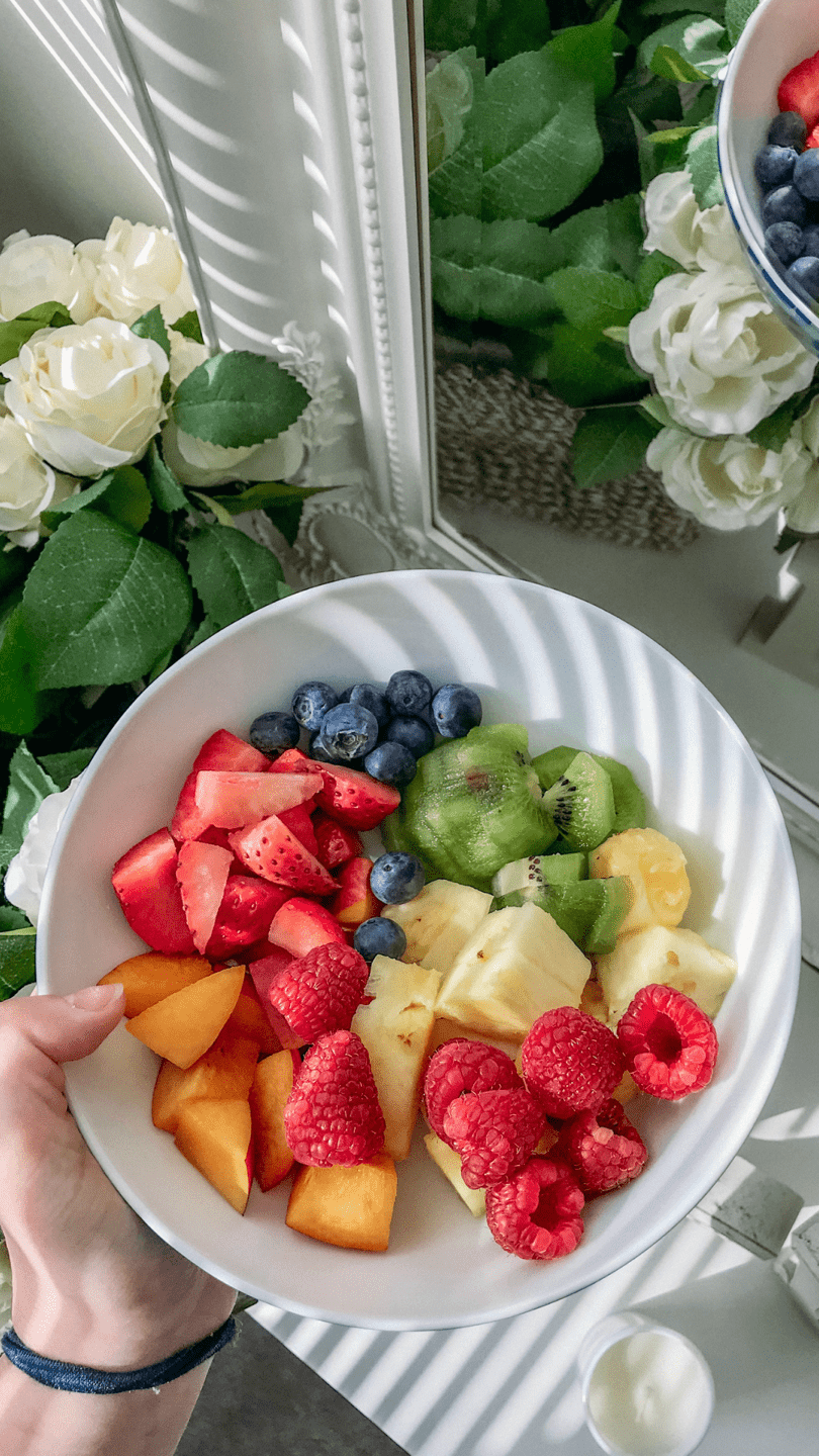 Enjoy the sweetness of life Background — A hand holds a white bowl filled with vibrant chopped strawberries raspberries blueberries kiwi and pineapple under bright window light.