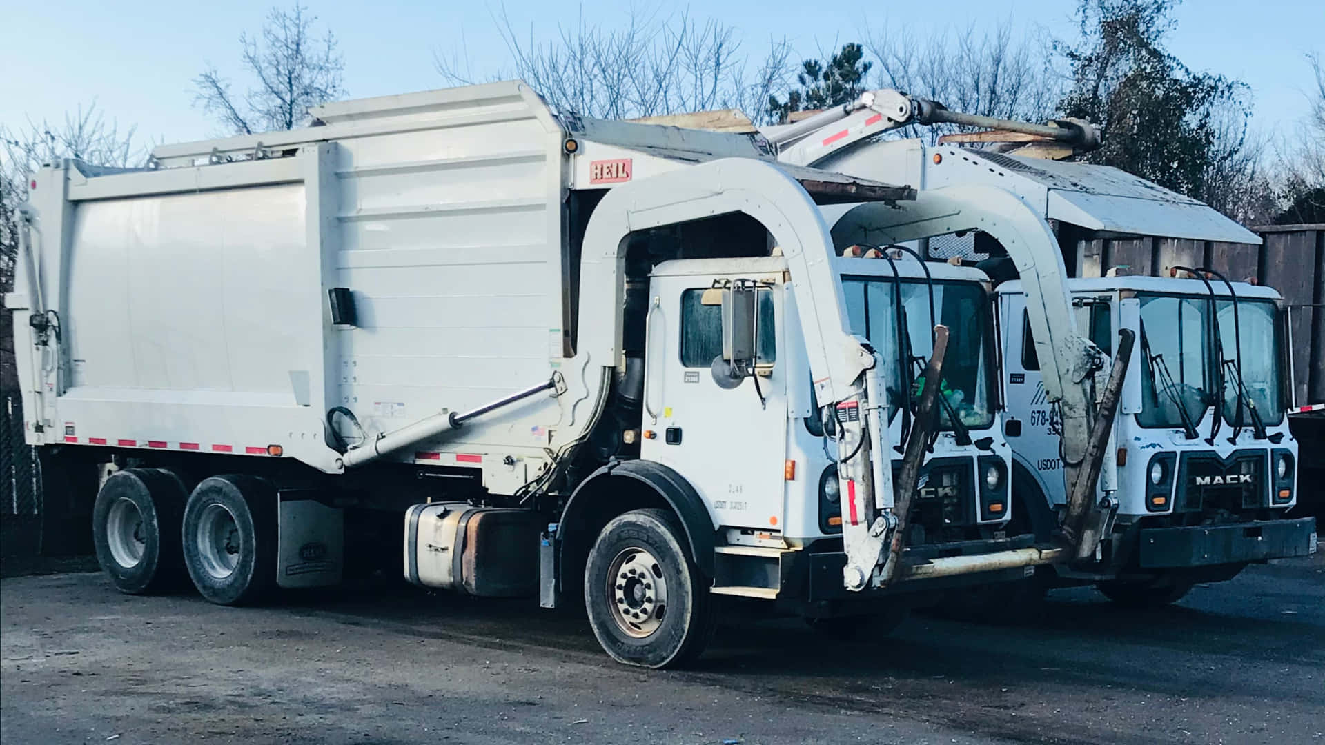 Two White Garbage Trucks Parked In A Parking Lot