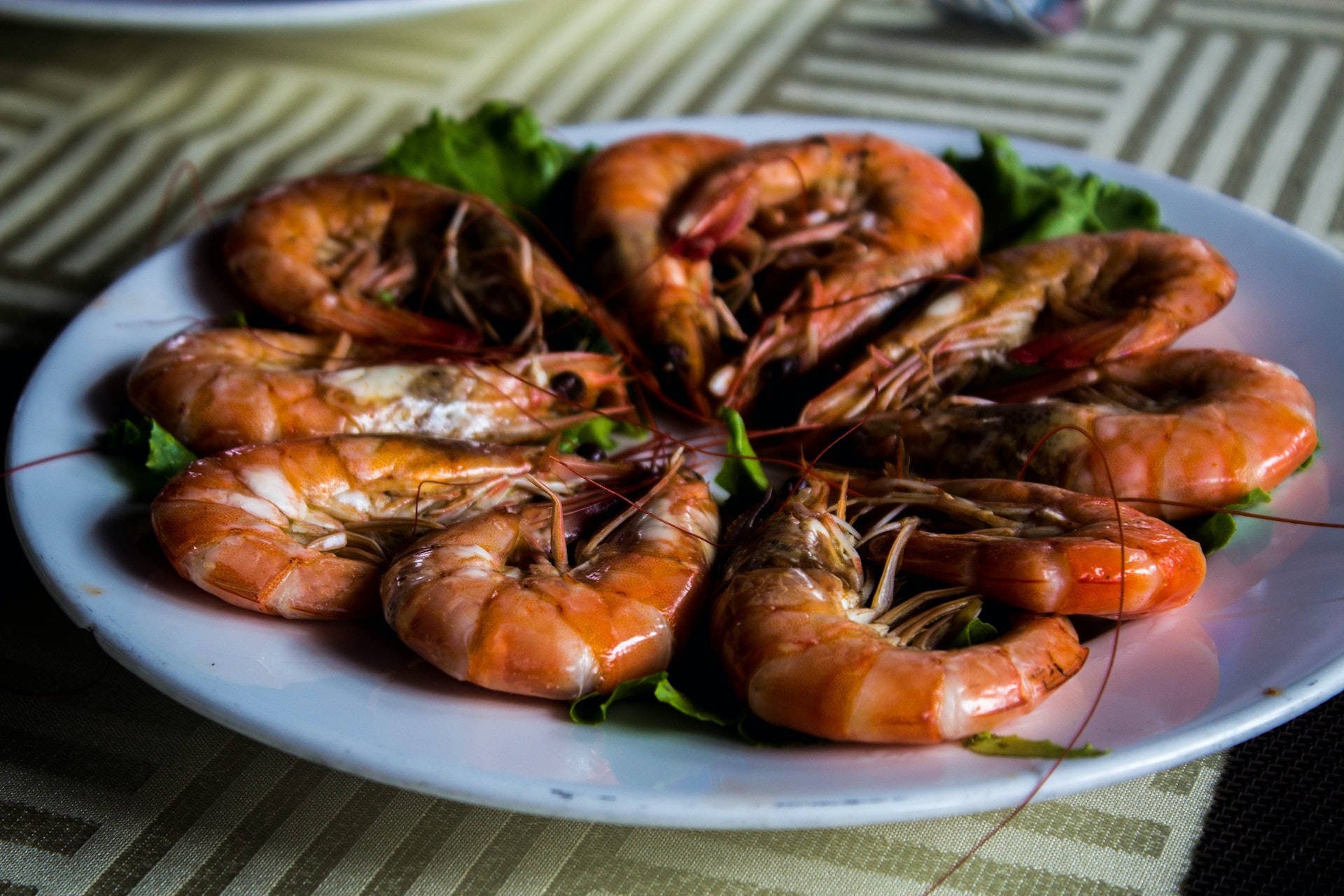 Prawns Sauté With Vegetables Wallpaper — A plate of glossy cooked shrimp arranged in a circle on green lettuce with a striped tablecloth in soft focus background.
