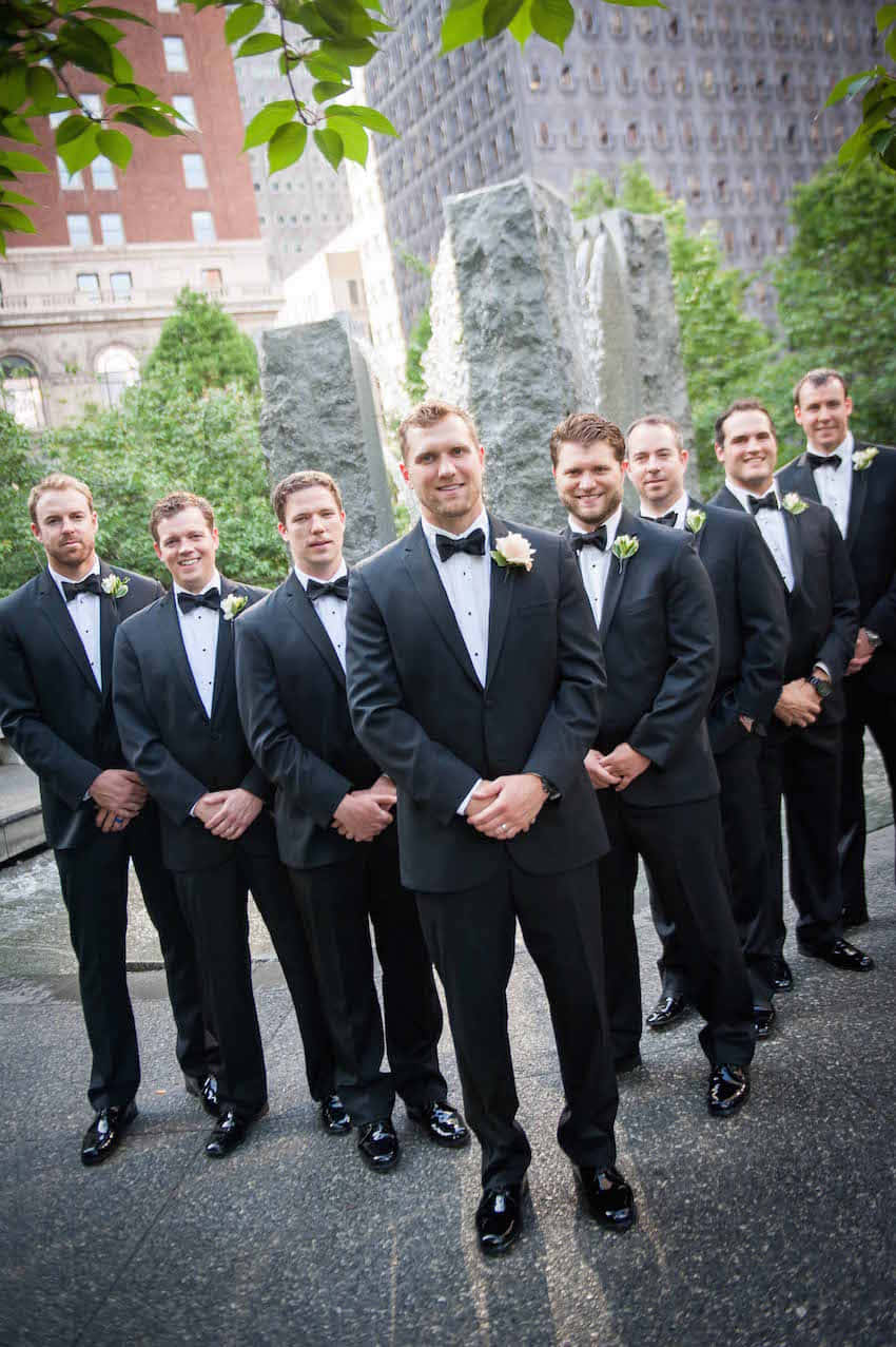 A Group Of Groomsmen In Tuxedos Standing In Front Of A Fountain