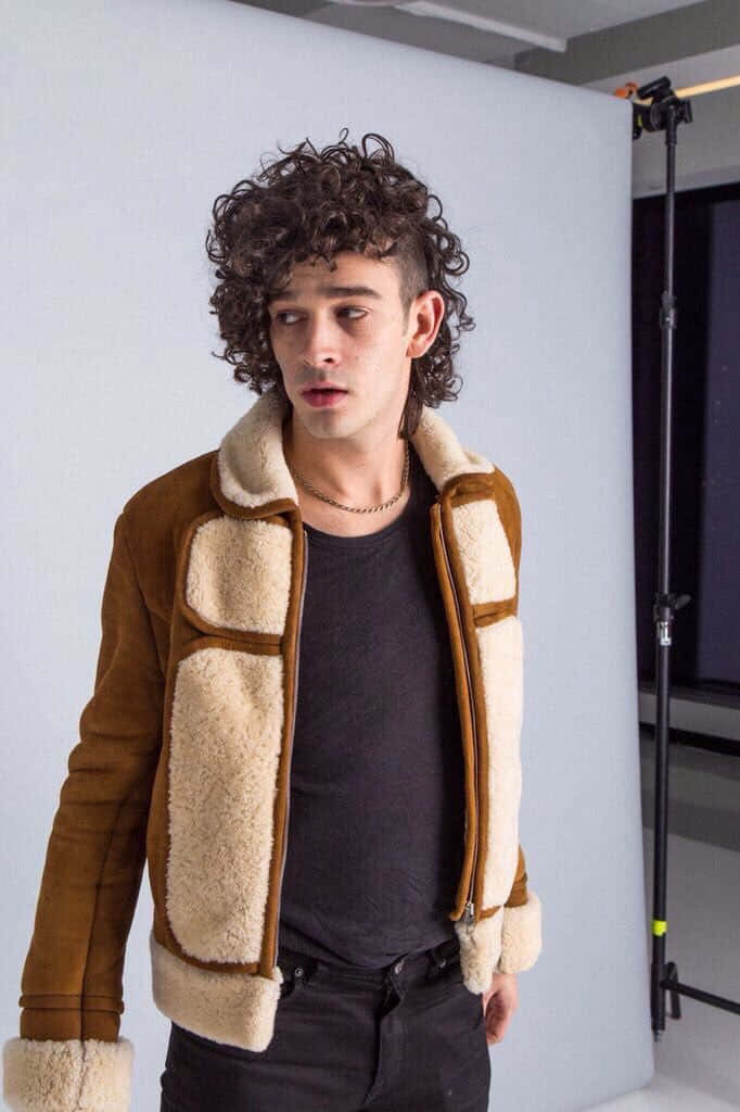 A Man In A Brown Shearling Jacket Standing In Front Of A Camera Wallpaper