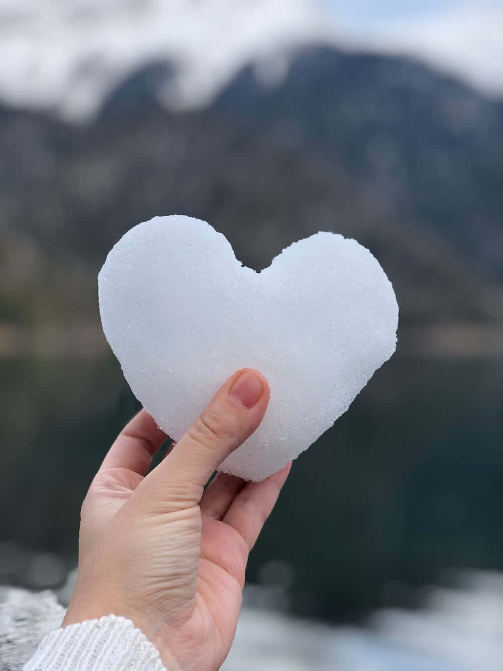 A Person Holding A Heart Shaped Piece Of Ice In Front Of A Lake