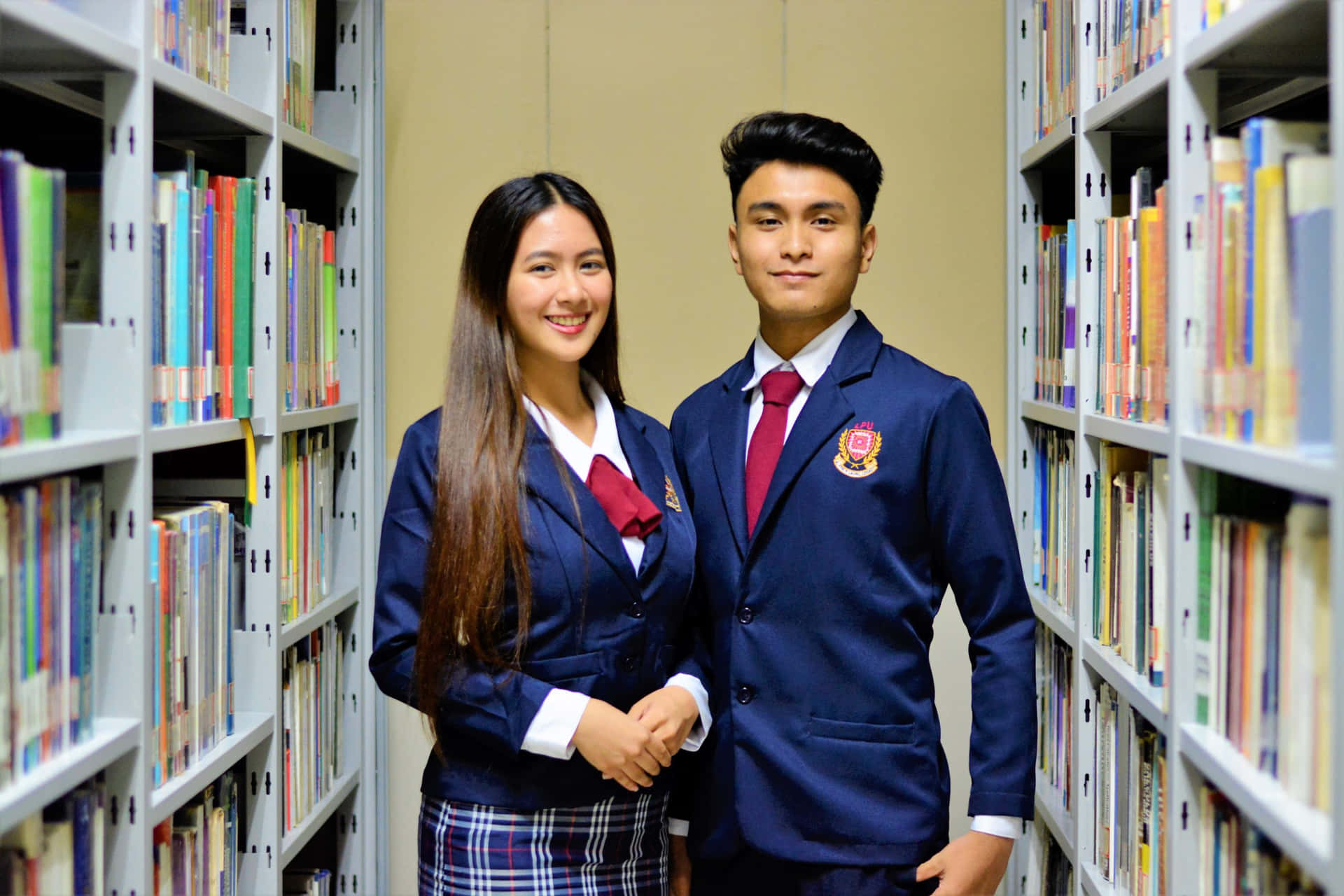 Two Young People In School Uniforms Standing In A Library