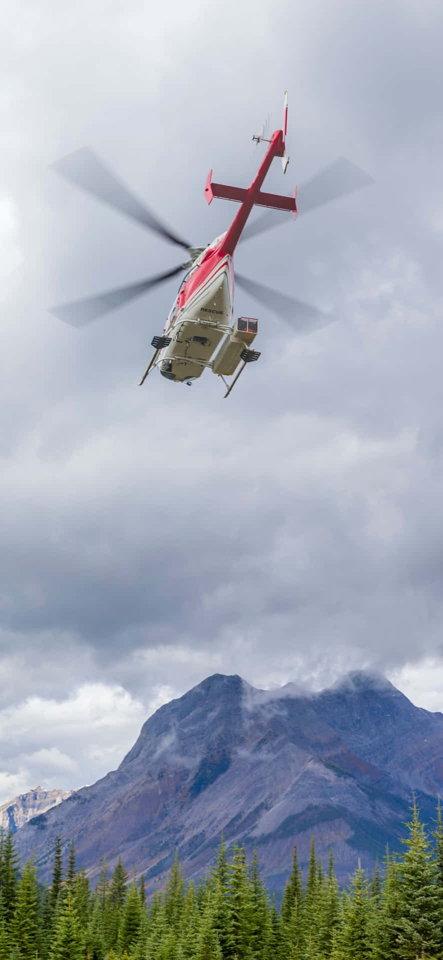 A Helicopter Flying Over A Forest With Mountains In The Background