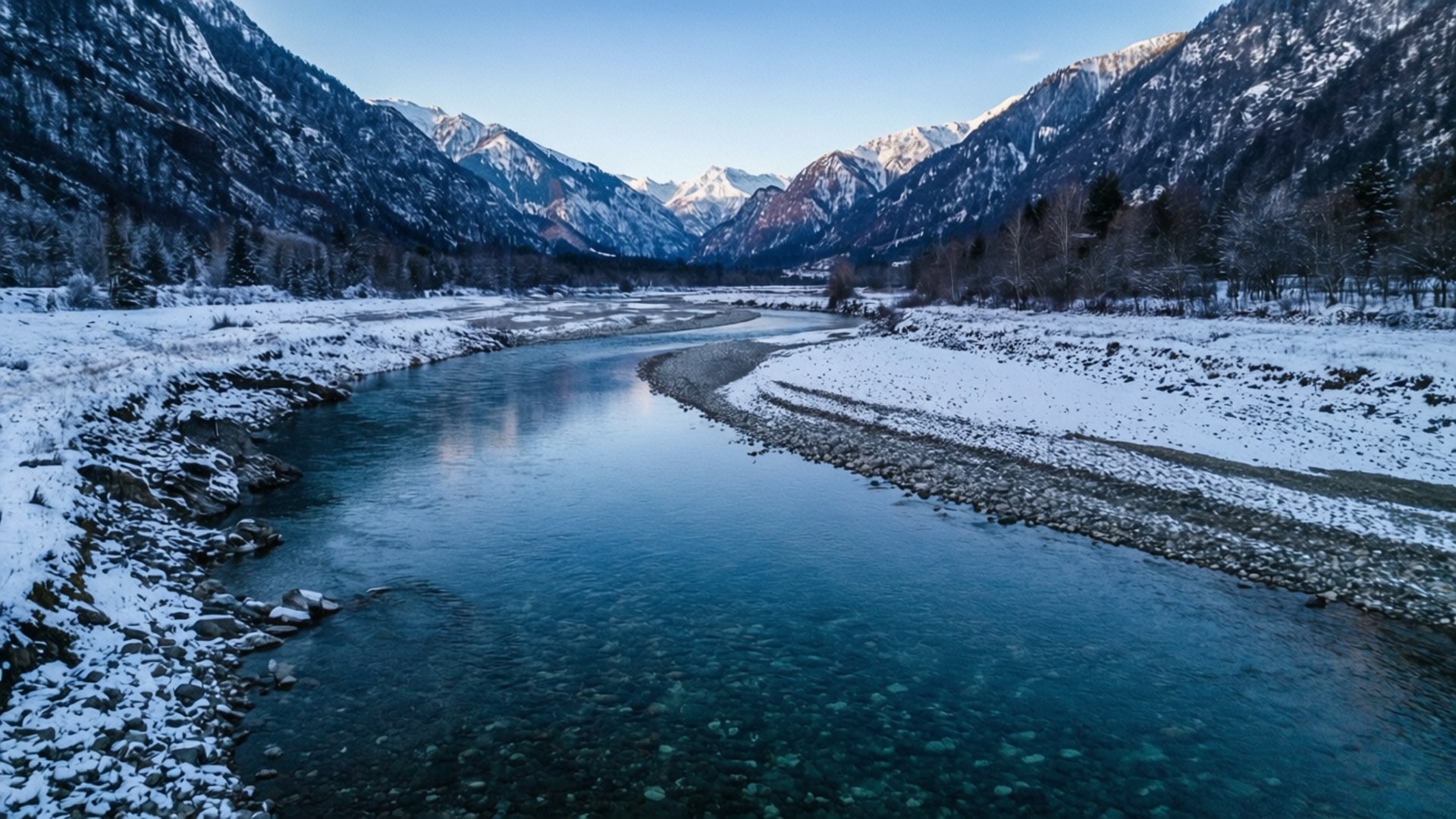 River Through Snow Wallpaper — A serene winter river winds through snow-covered mountains under a clear blue sky.