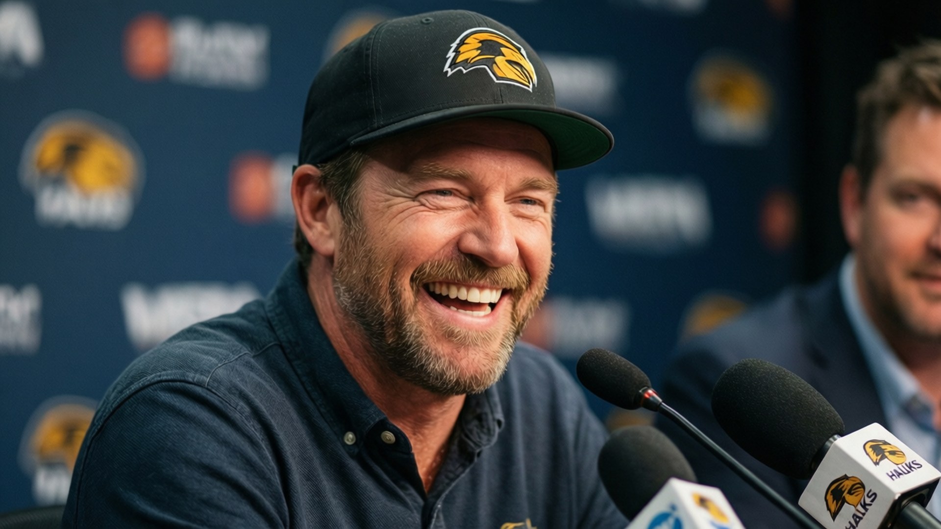Laughing Interview Wallpaper — A smiling man in a black hawk logo cap laughs at a press conference with microphones and branded backdrop.
