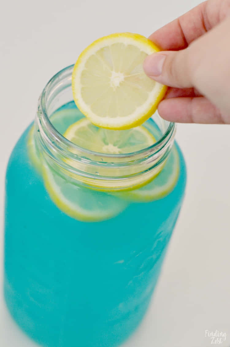 A Person Is Putting Lemon Slices Into A Jar Of Blue Liquid