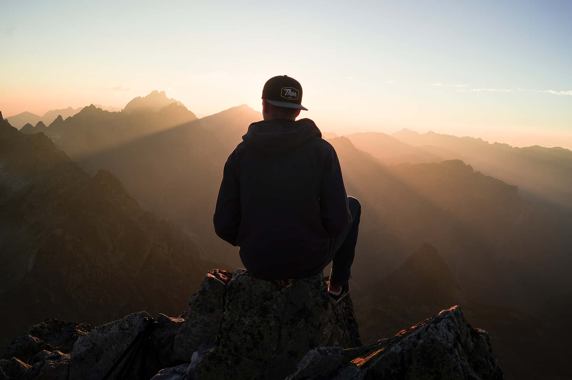 A Man Sitting On Top Of A Mountain At Sunset