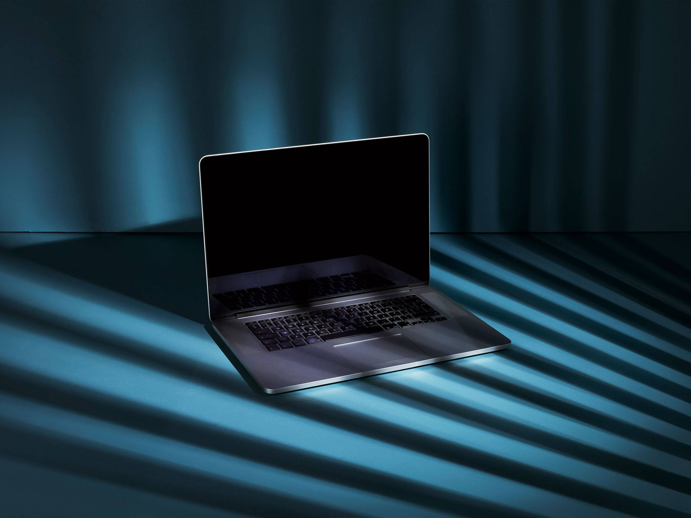 MacBook With Webcam Wallpaper — A sleek silver laptop rests on a floor bathed in diagonal blue light casting sharp shadows against a draped curtain backdrop.