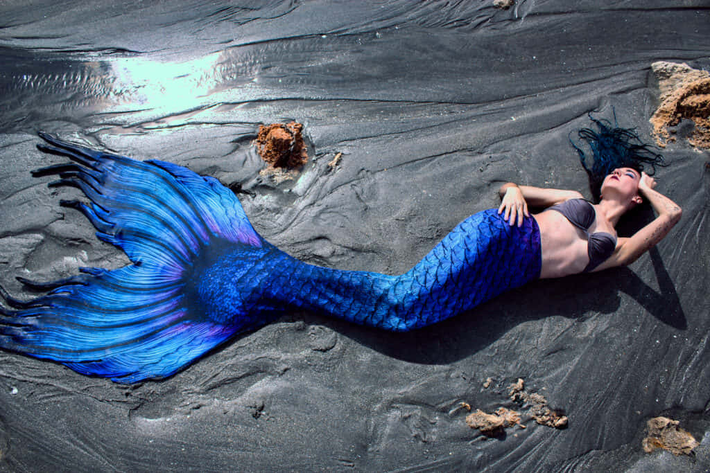 a woman laying on the beach with a blue mermaid tail