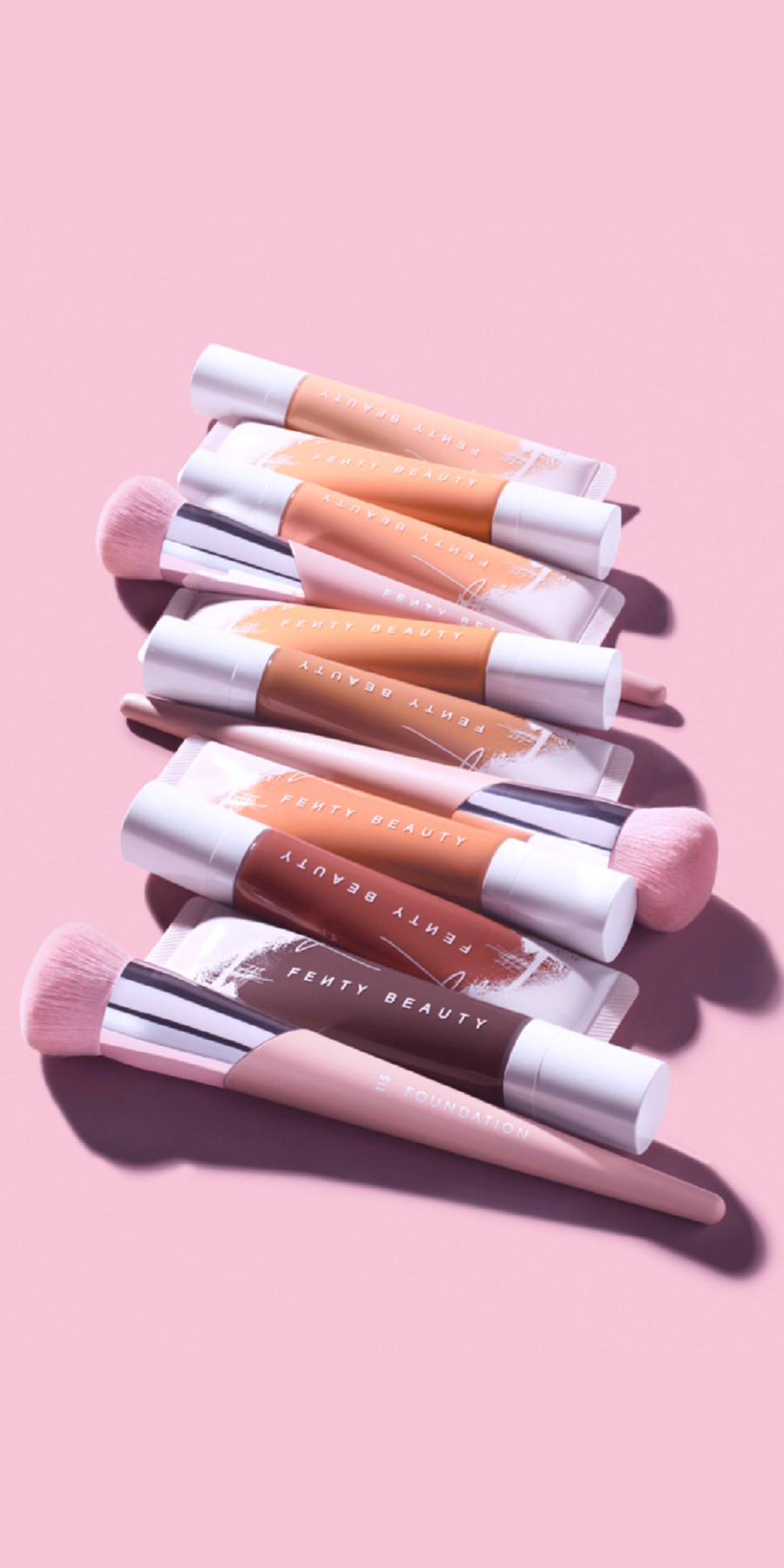 Makeup Background — A diagonal arrangement of Fenty Beauty lip gloss tubes and brushes on a soft pink background with sharp shadows.
