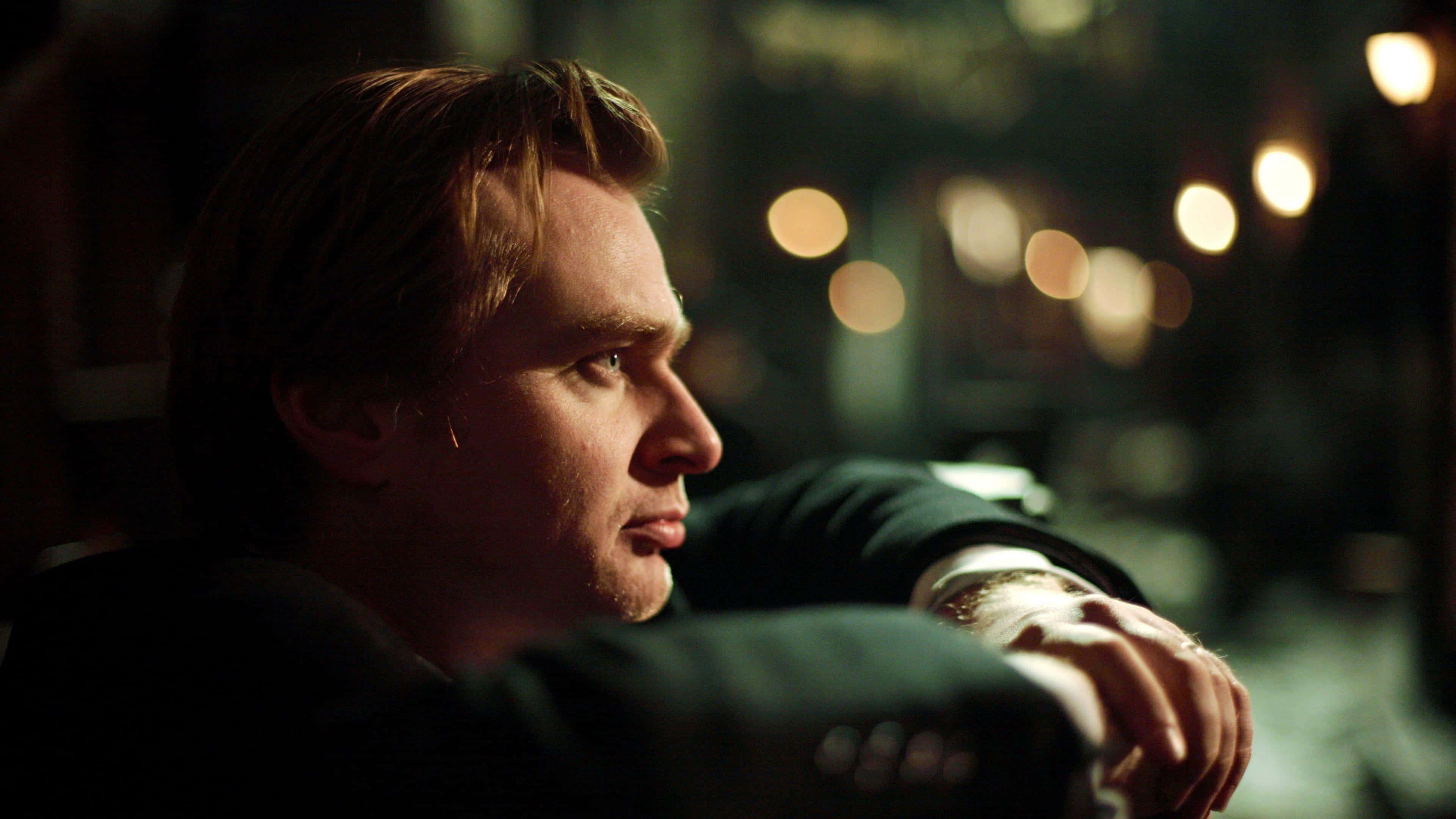 Mastermind Director, Christopher Nolan Wallpaper — A man in a dark suit gazes thoughtfully out a window at blurred city lights glowing warmly in the night.