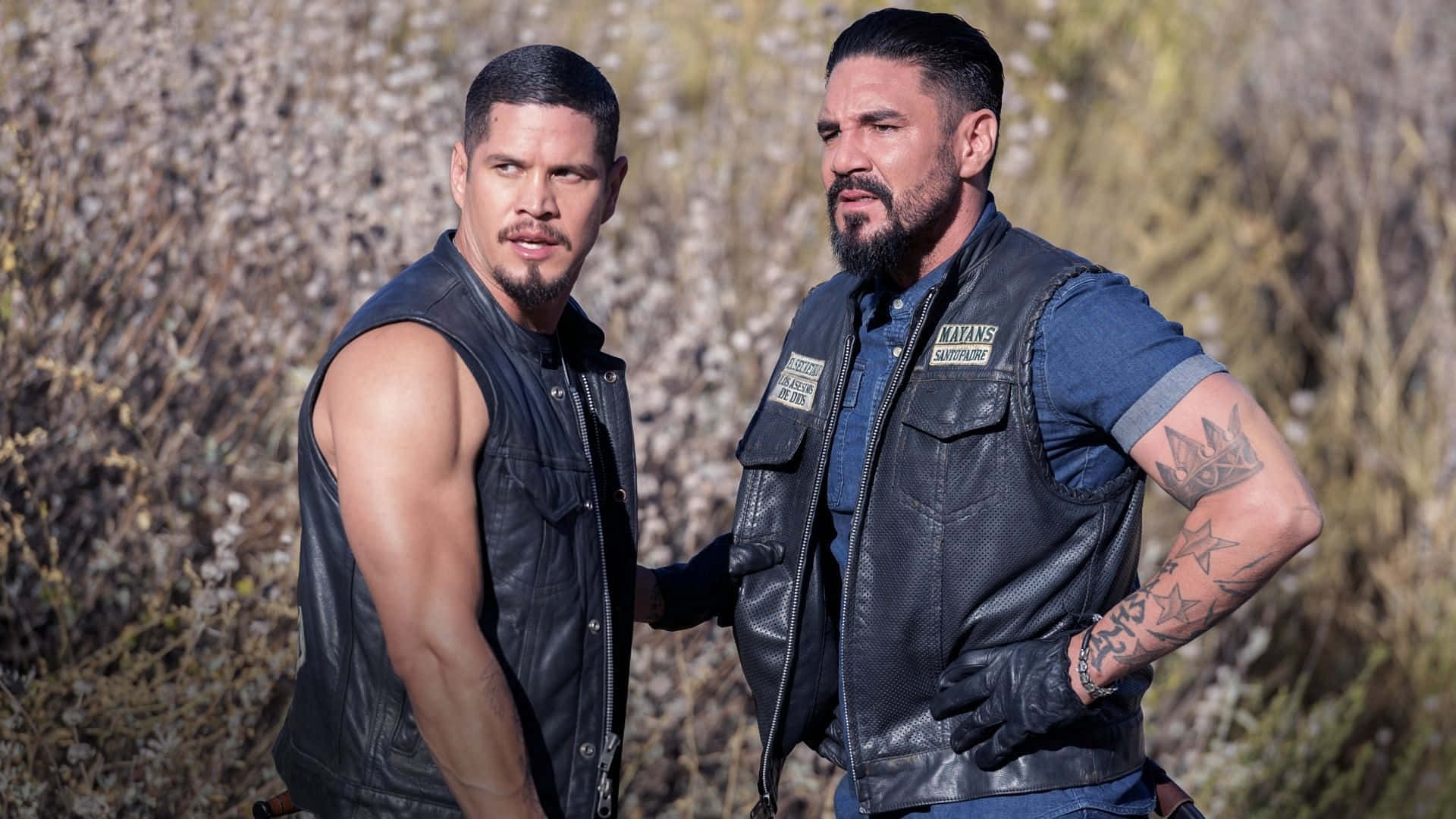 Two Men In Leather Vests Standing In A Field Wallpaper