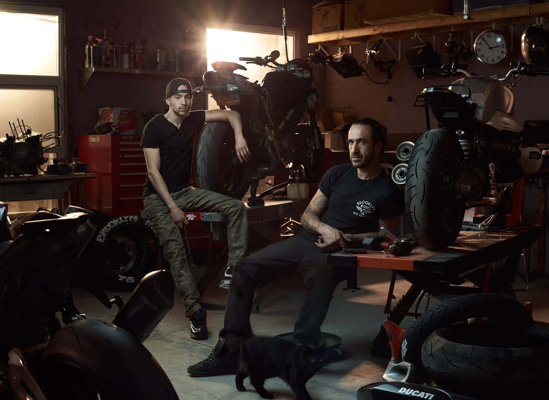 Two Men Sitting In A Garage With Motorcycles