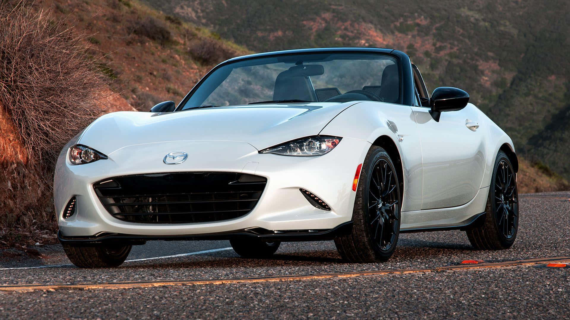 The White Mazda Mx-5 Roadster Is Driving Down A Mountain Road Wallpaper