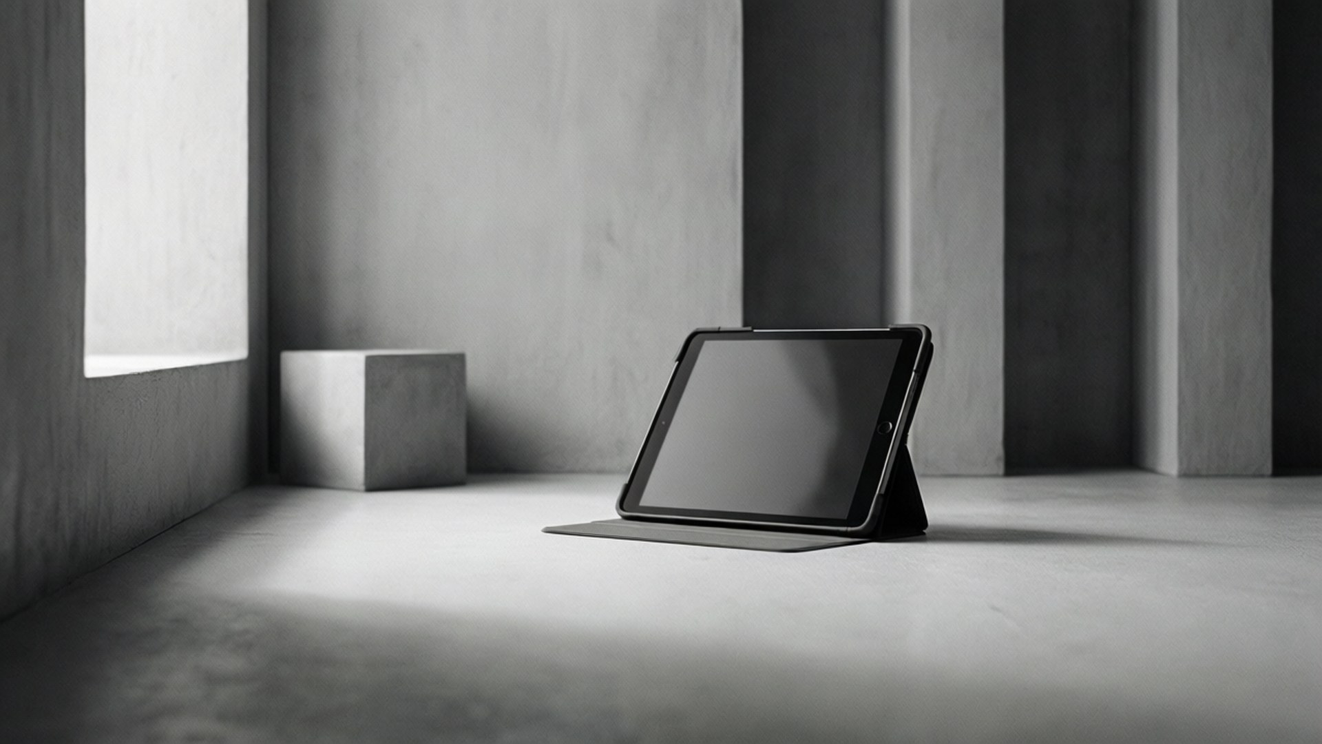 Minimalist Tablet Wallpaper — A sleek black tablet rests on a minimalist concrete floor in a stark monochrome room with geometric pillars and soft natural light.