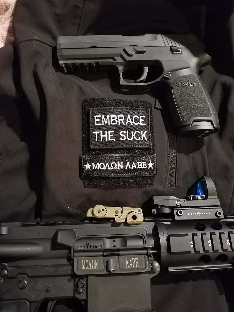 A Gun And A Patch With The Words Embrace The Suck Wallpaper