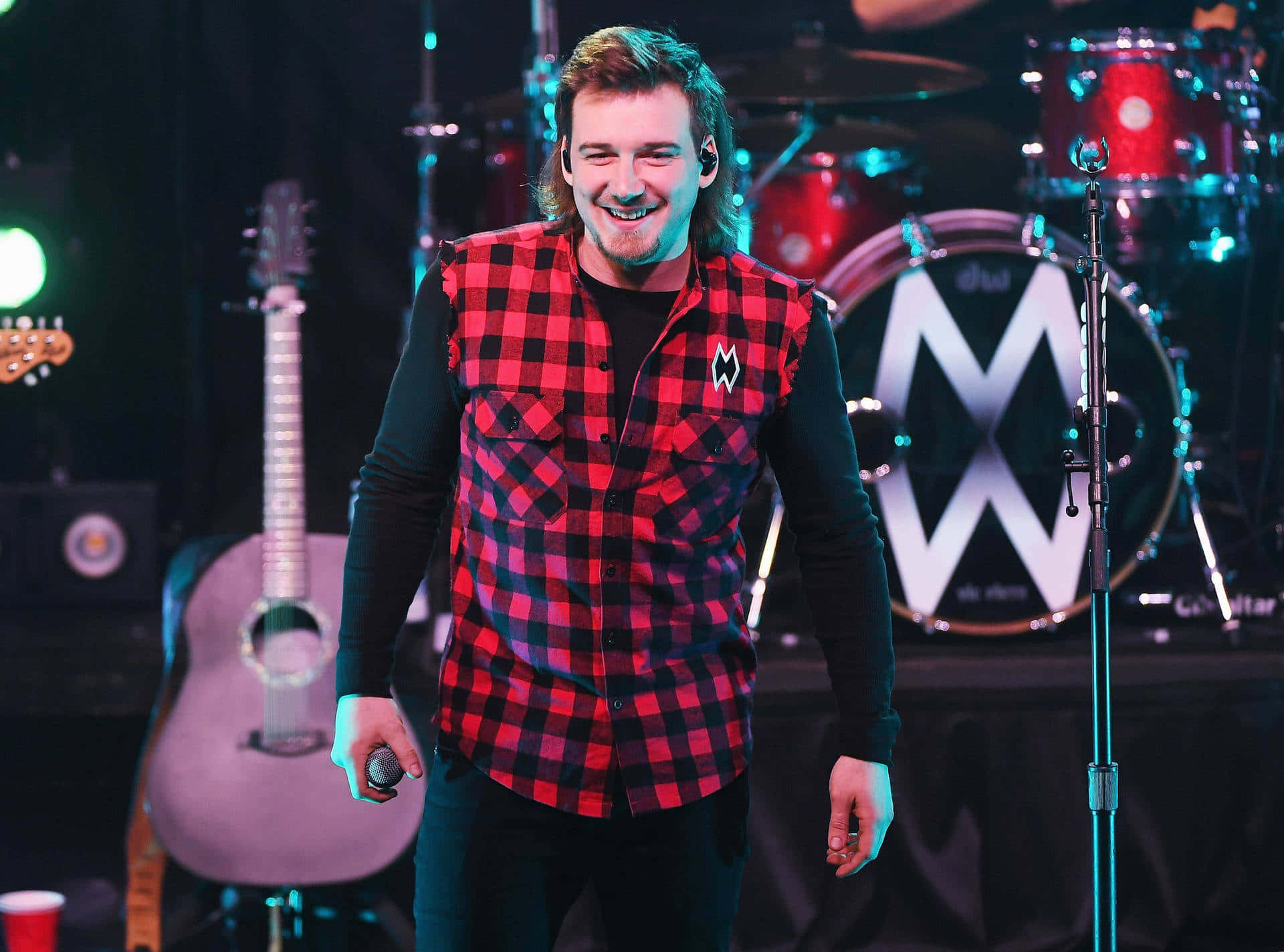 A Man In A Plaid Shirt Is Smiling On Stage