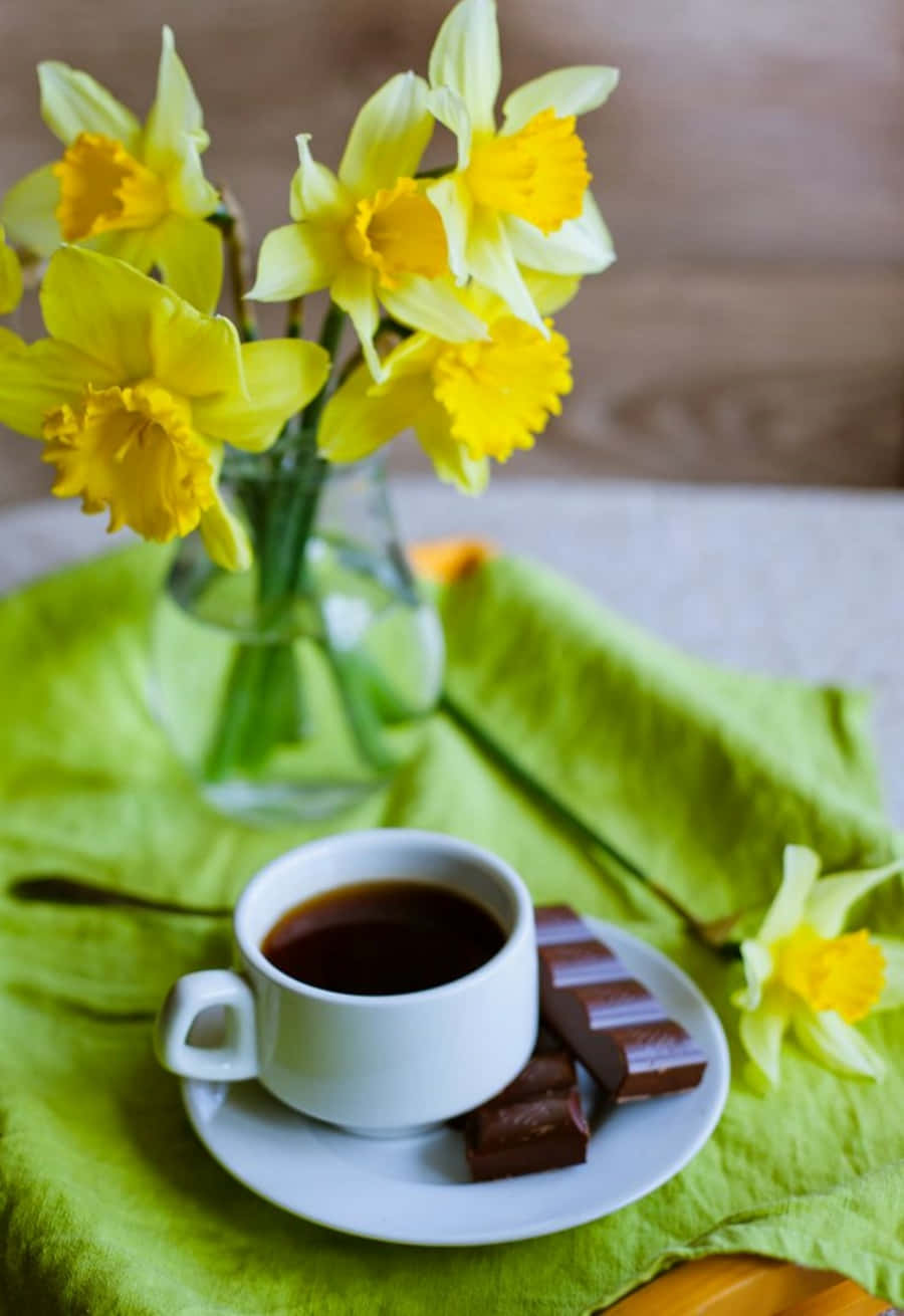 A Cup Of Coffee With Daffodils And Chocolate On A Green Cloth
