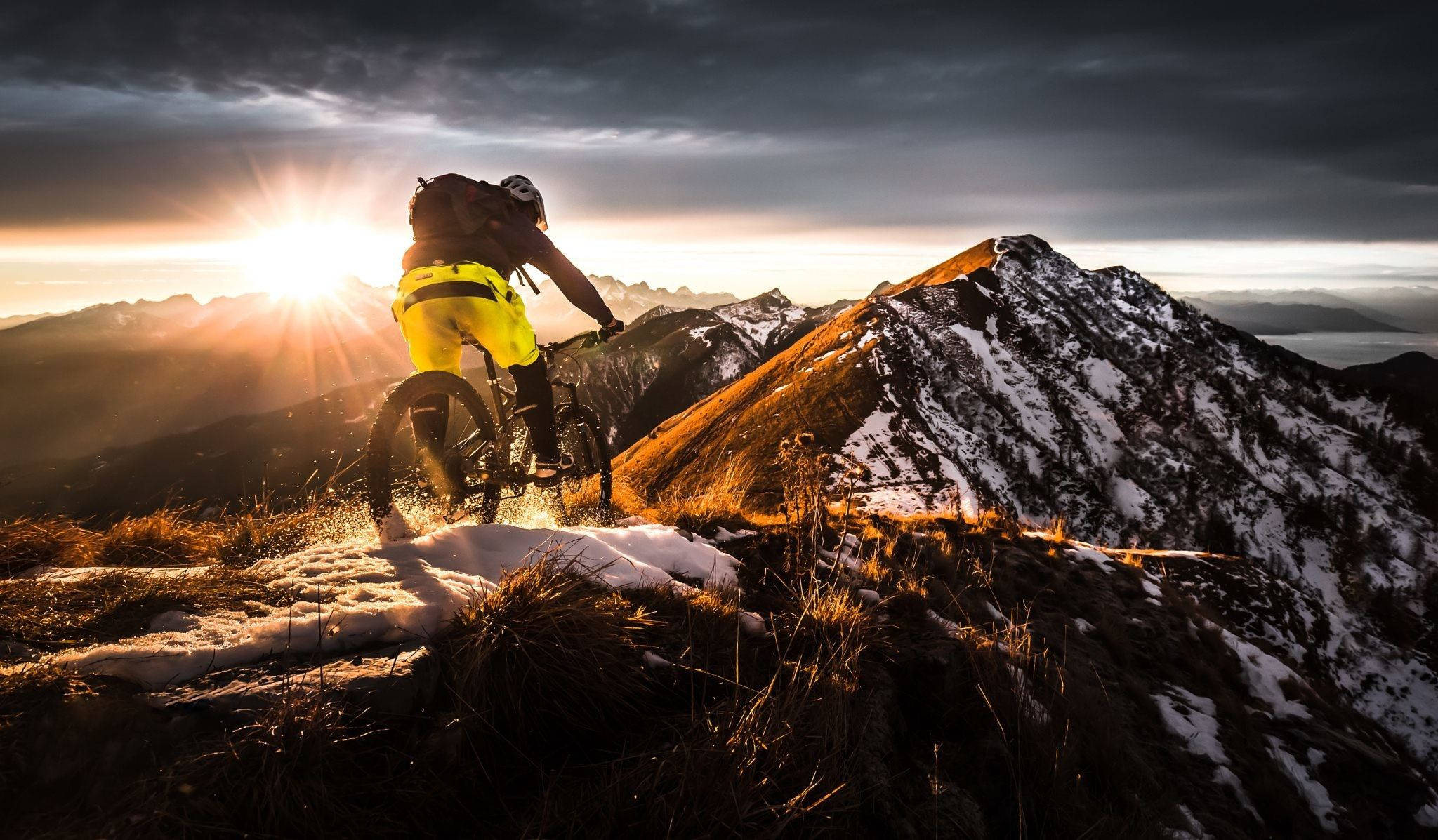 Thrilling MTB Ride on a Snow-Covered Mountain Wallpaper — A mountain biker in bright yellow pants rides through snow at sunset on a rugged alpine ridge with dramatic clouds.