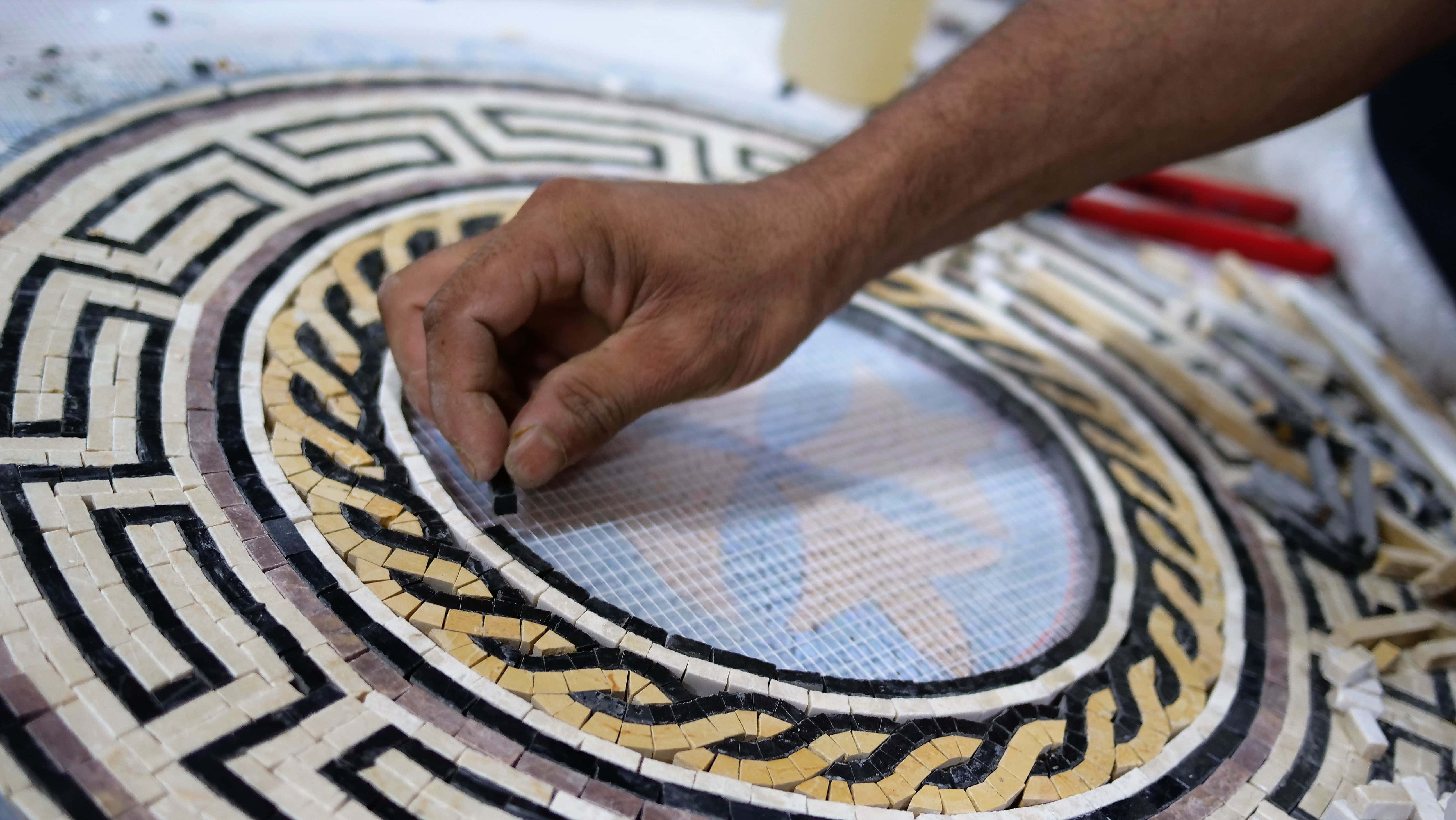 Multi-Colored Mosaic Patterns Picture — A craftsman's hand places a tiny black tile into a circular geometric mosaic with intricate black beige and white patterns.