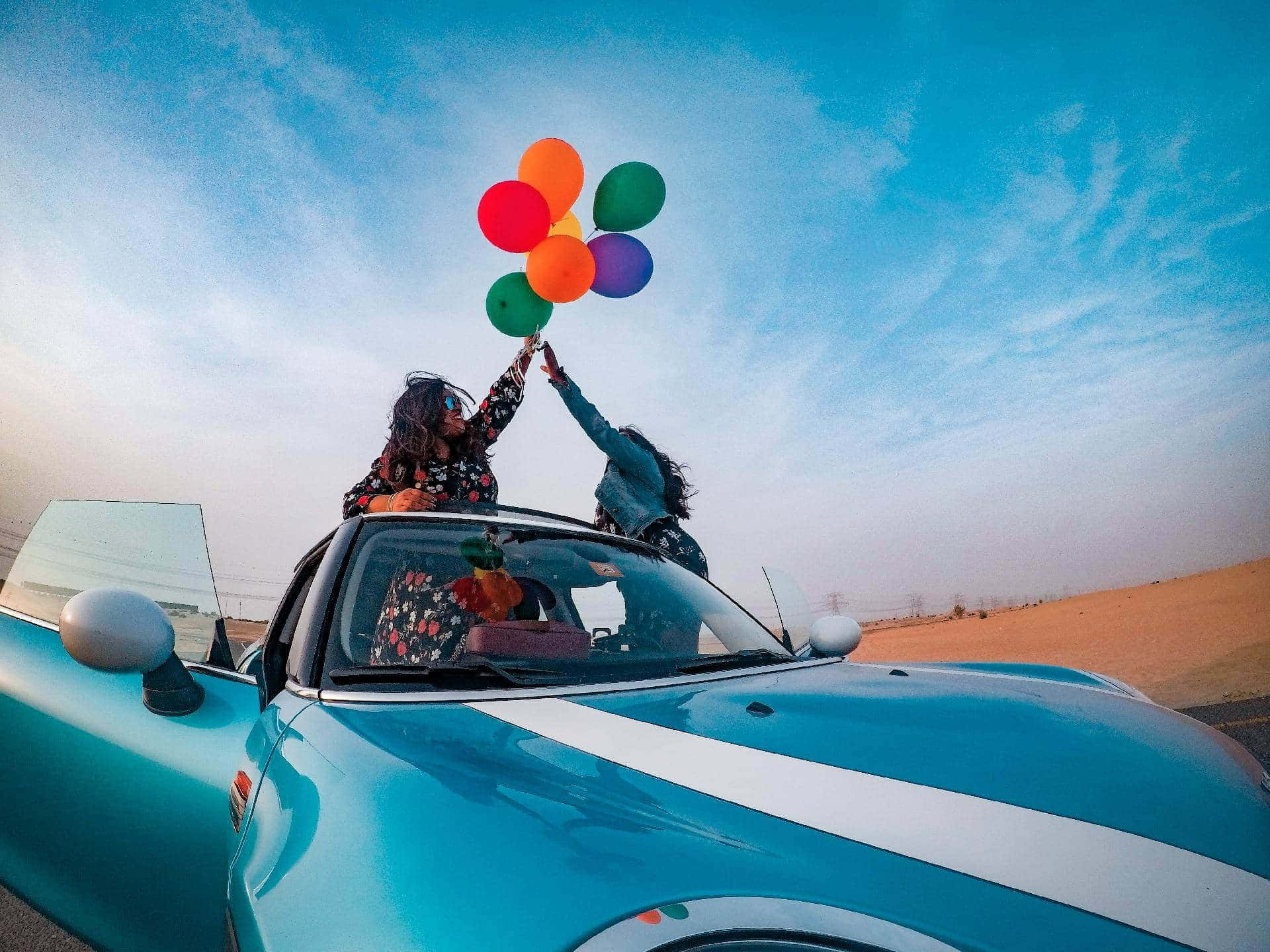 Two Women In A Blue Convertible Car Holding Balloons