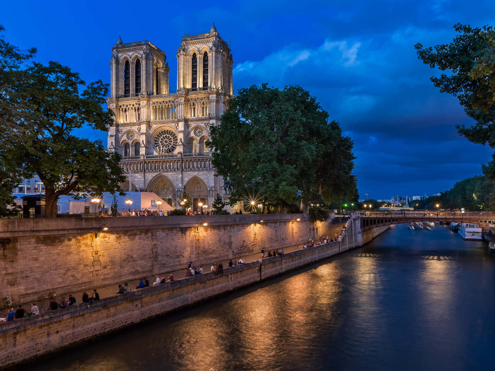 Notre Dame Cathedral With Tourists On The River Pathway Wallpaper