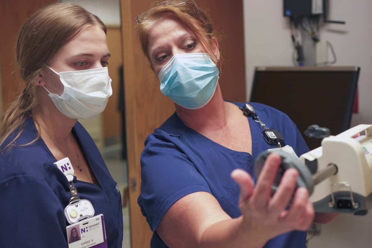Two Nurses In Scrubs Are Looking At A Machine