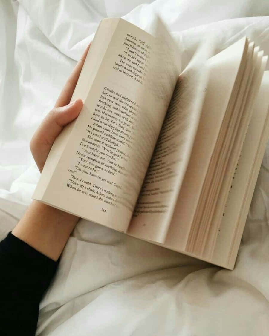 A Person Holding An Open Book On A Bed