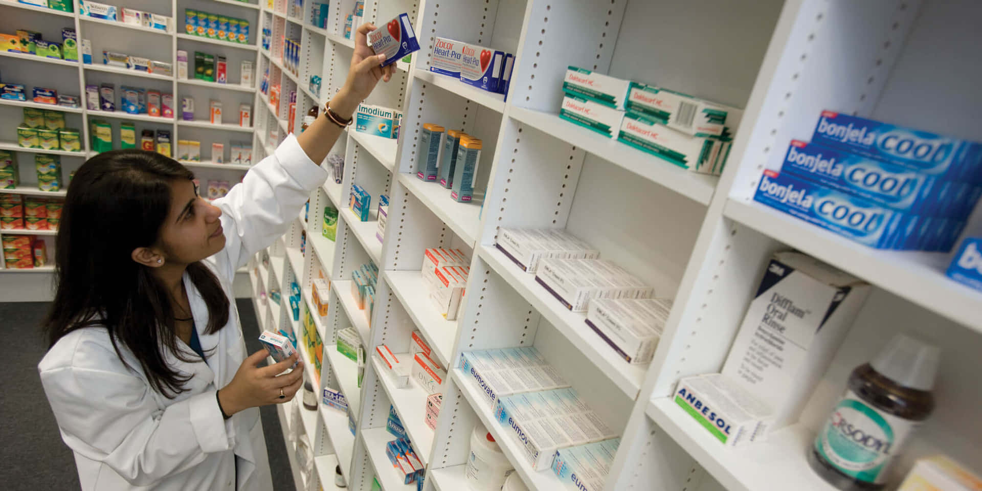 A Woman In A White Coat Is Looking At Shelves Of Medicine