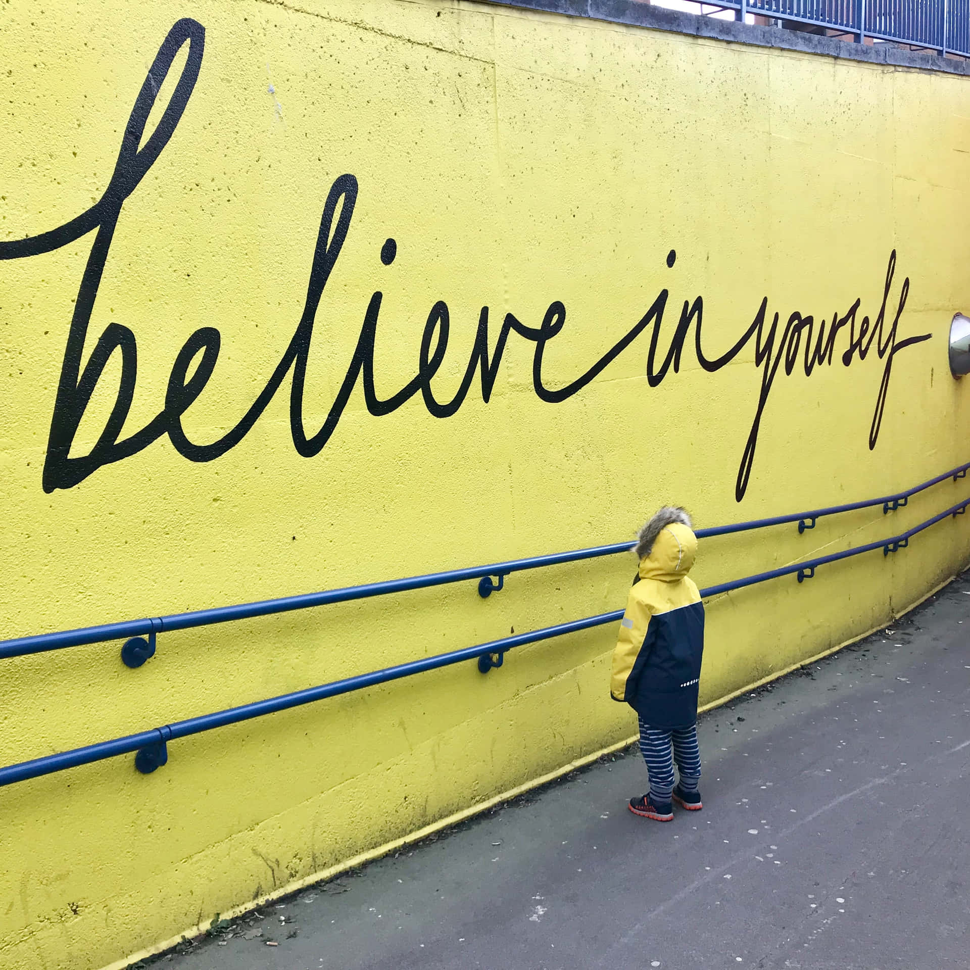 Et barn står ved siden af ​​en gul væg med et citat, der siger tro på dig selv. Wallpaper